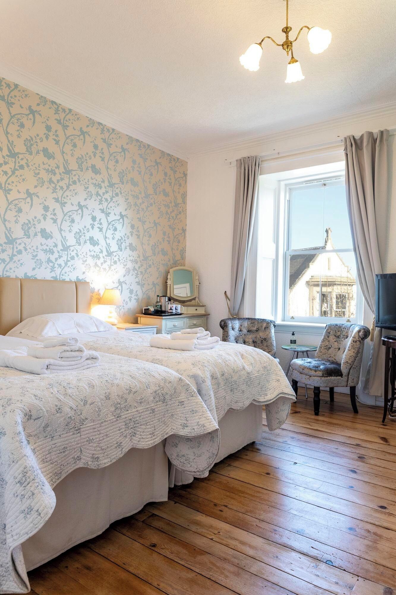 Habitación de hotel con una cama grande, una mesita de noche, un espejo, una silla, un televisor y una ventana con cortinas.
