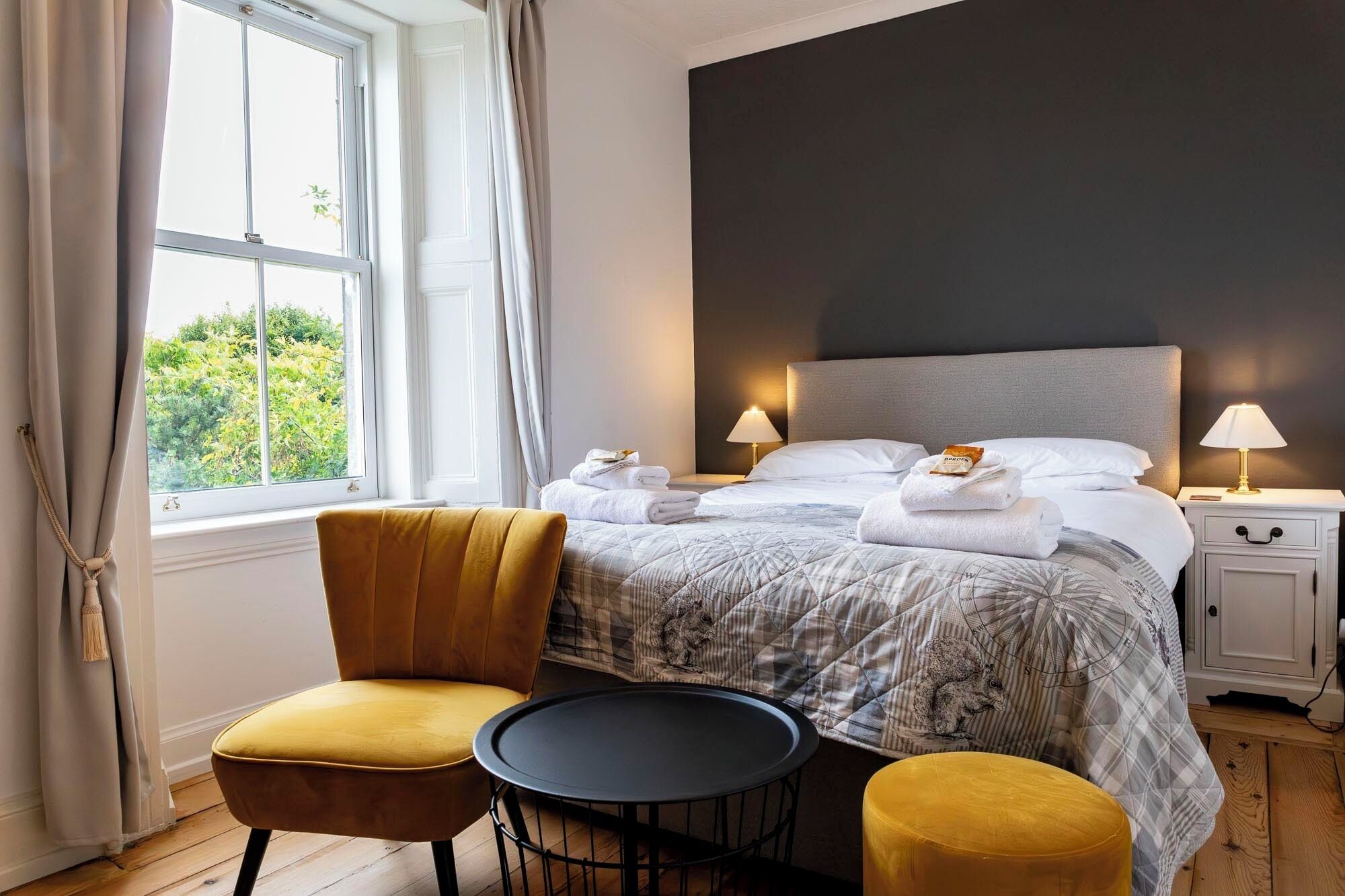 A hotel room with a bed, two bedside lamps, a chair, a small table, and a window with curtains.