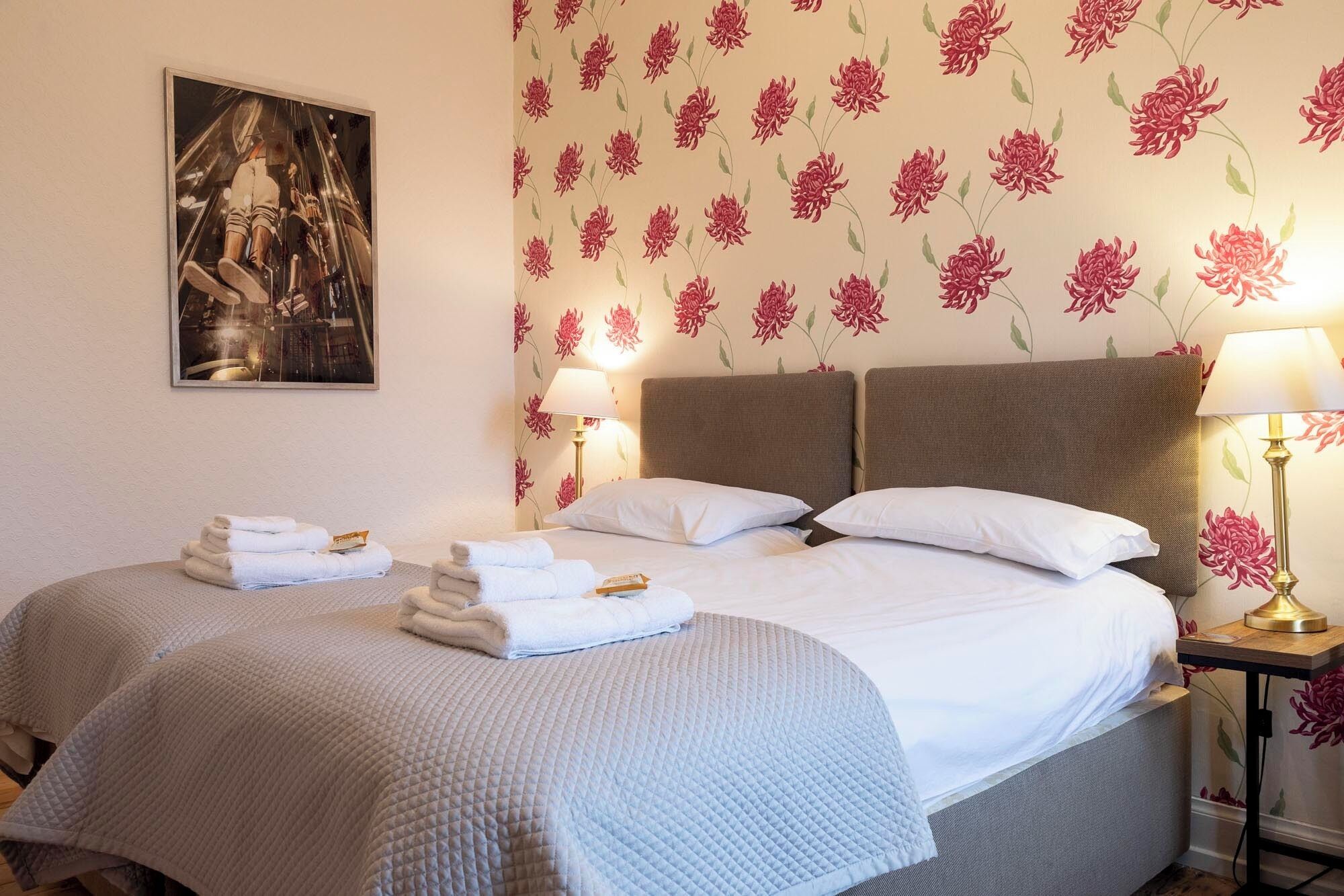 Een hotelkamer met twee bedden, een schilderij aan de muur, bloemenbehang en een nachtkastje met een lamp.