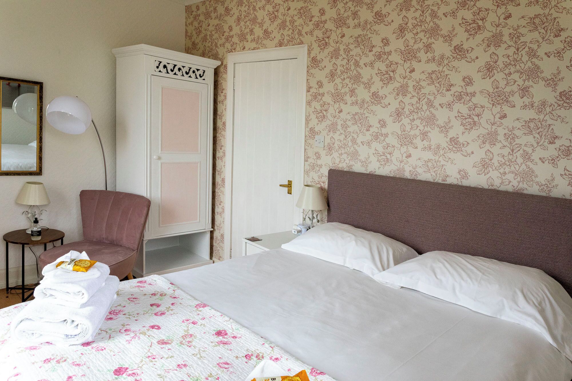 A hotel room with a bed, a chair, a wardrobe, and a nightstand with a lamp.