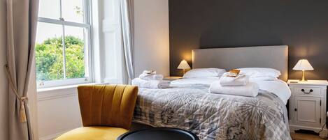 A hotel room with a bed, two bedside lamps, a chair, a small round table, and a window with curtains.