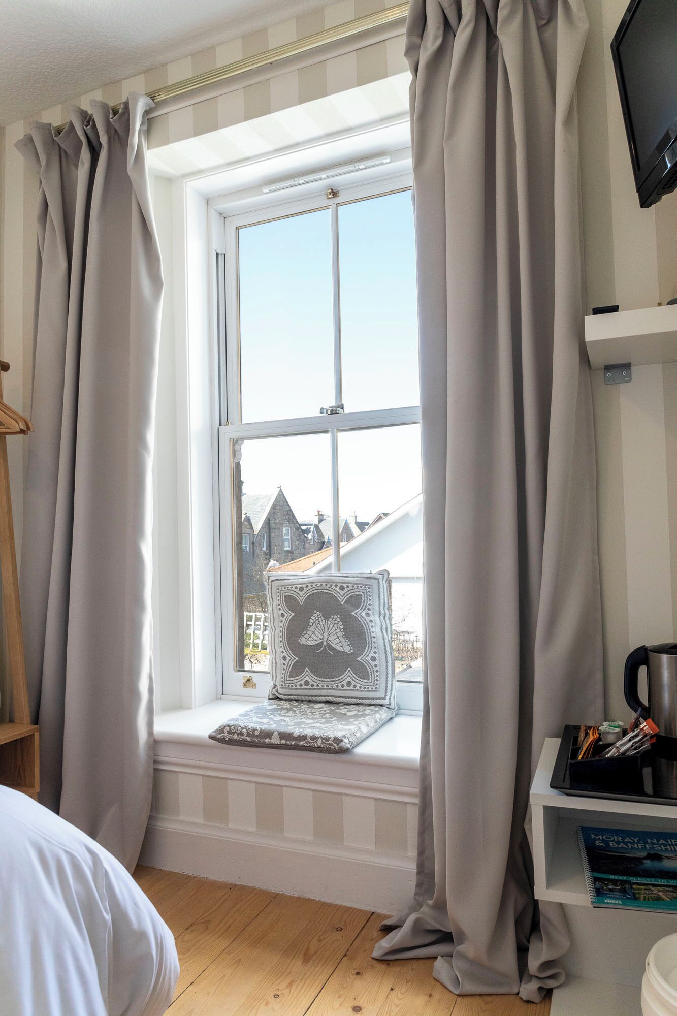 A room with a window, a bed, a bedside table, and a shelf with books.
