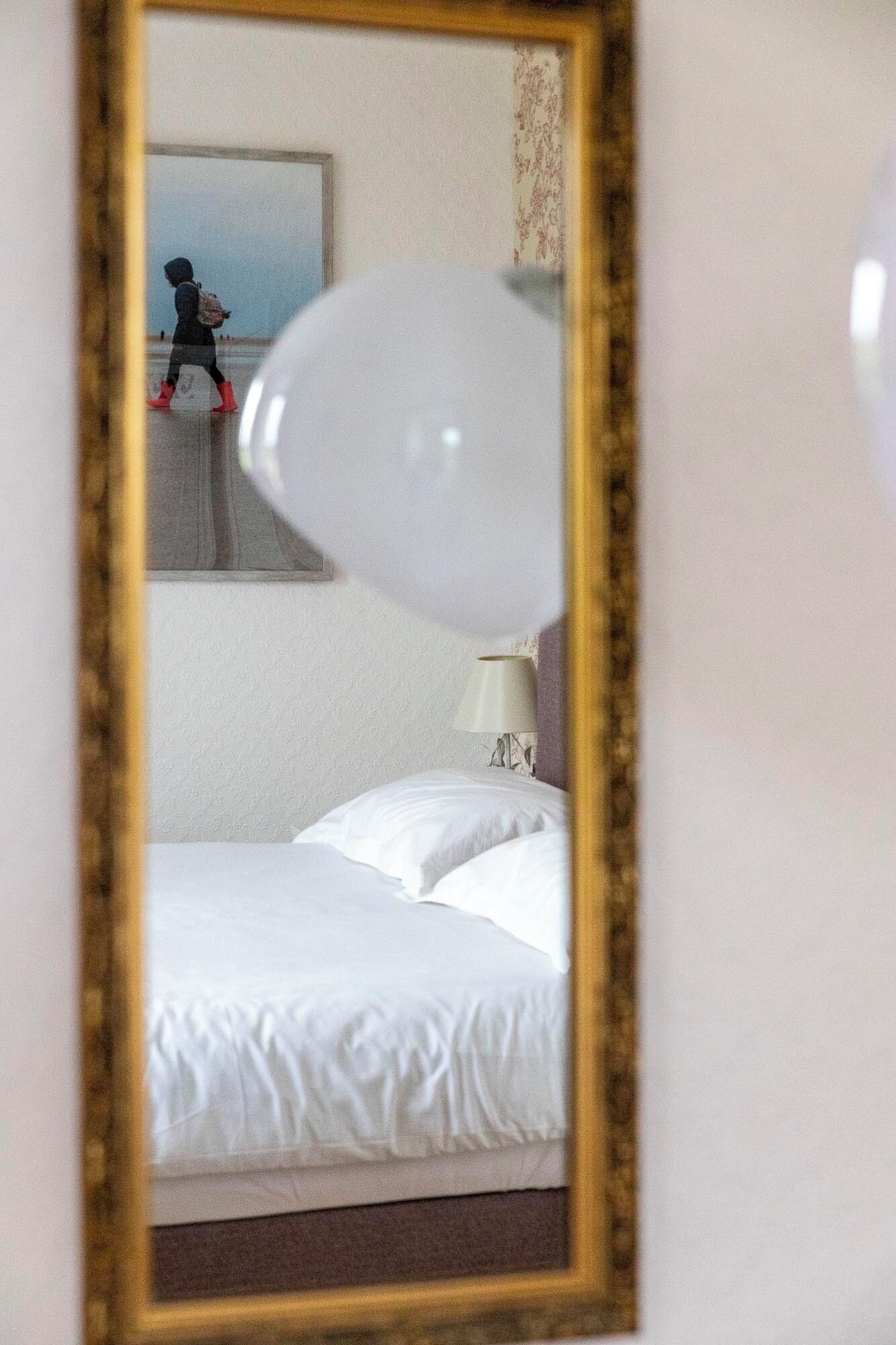A hotel room with a bed, a lamp, and a mirror reflecting a person walking outside.
