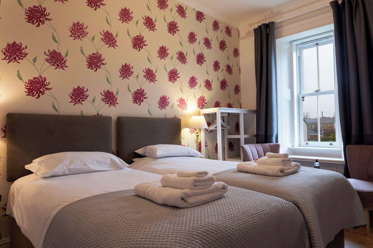 Camera d'albergo con due letti, una finestra con tende, un comodino con una lampada e una sedia.