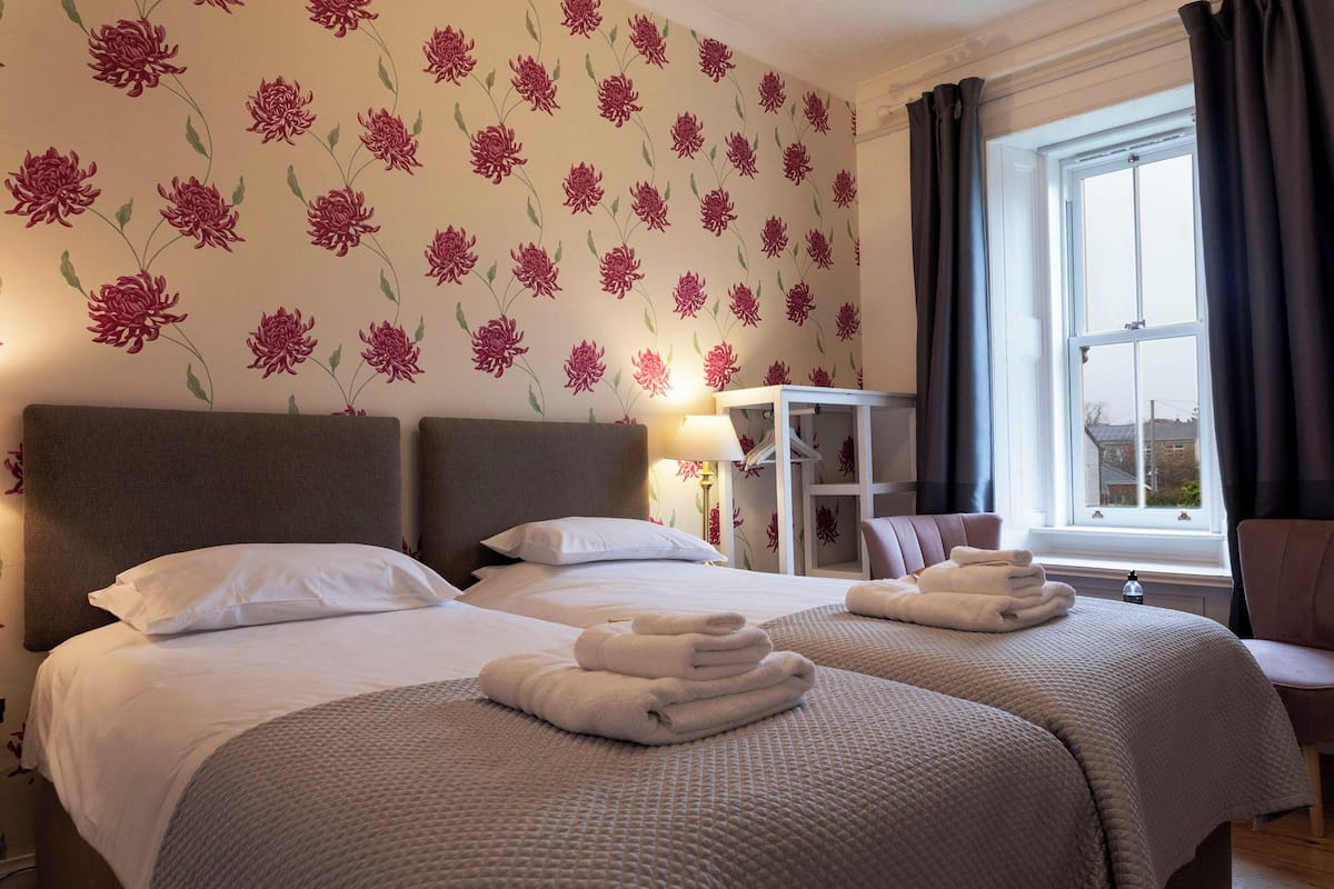 A hotel room with two beds, a window with curtains, a bedside table with a lamp, and a chair.