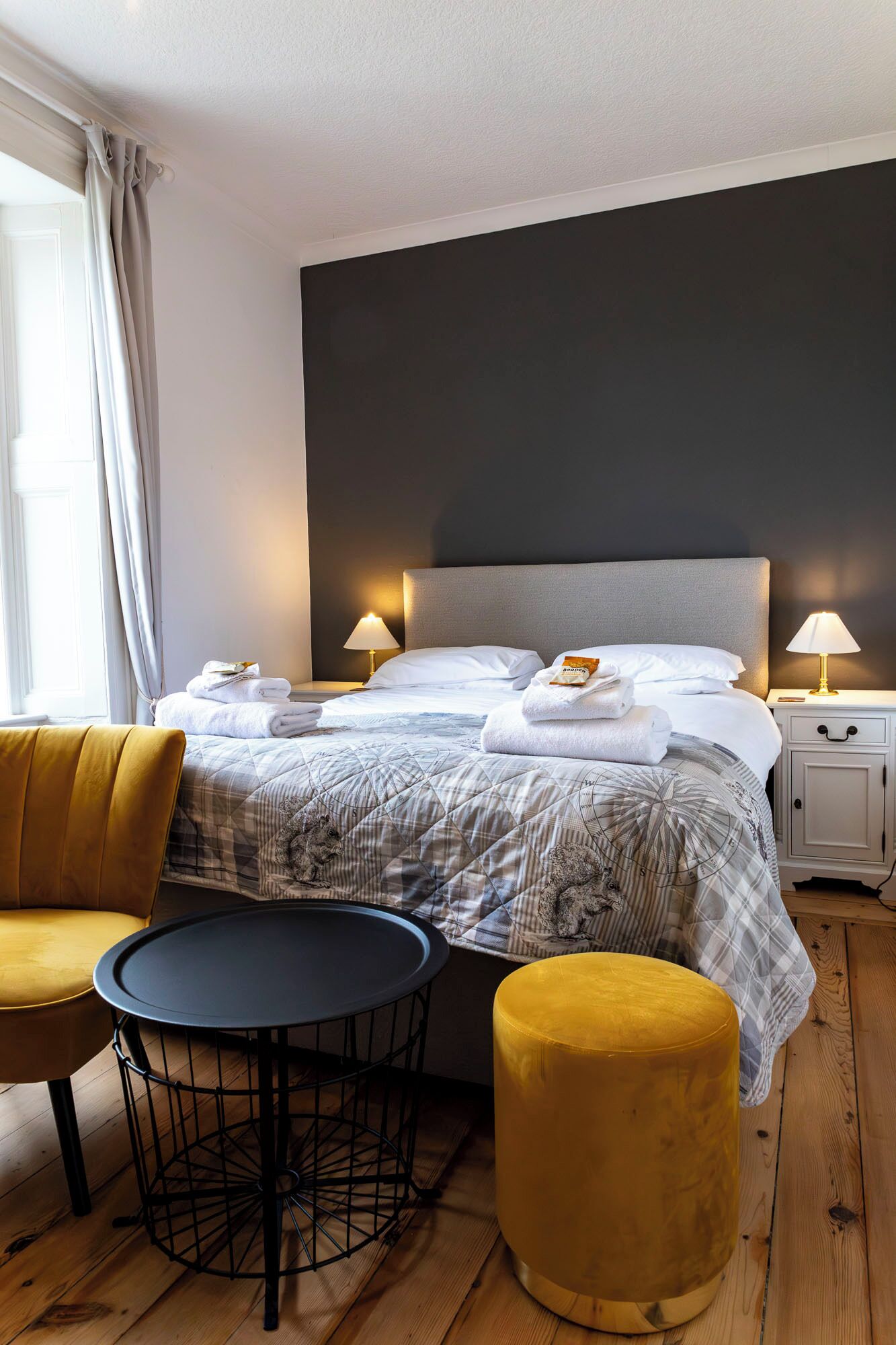 A hotel room with a bed, bedside tables, a round table, a yellow armchair, and a round ottoman.
