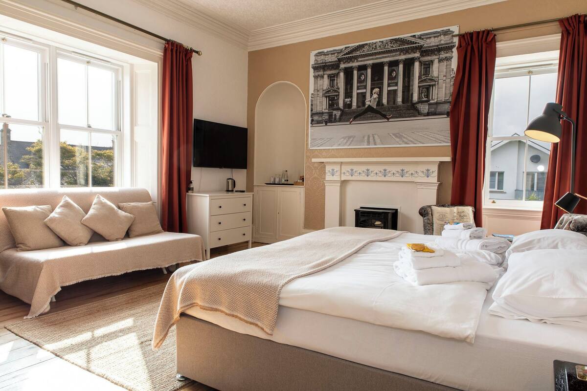 Una camera d'albergo con un letto, un divano, un camino, una televisione e un'ampia finestra con tende.