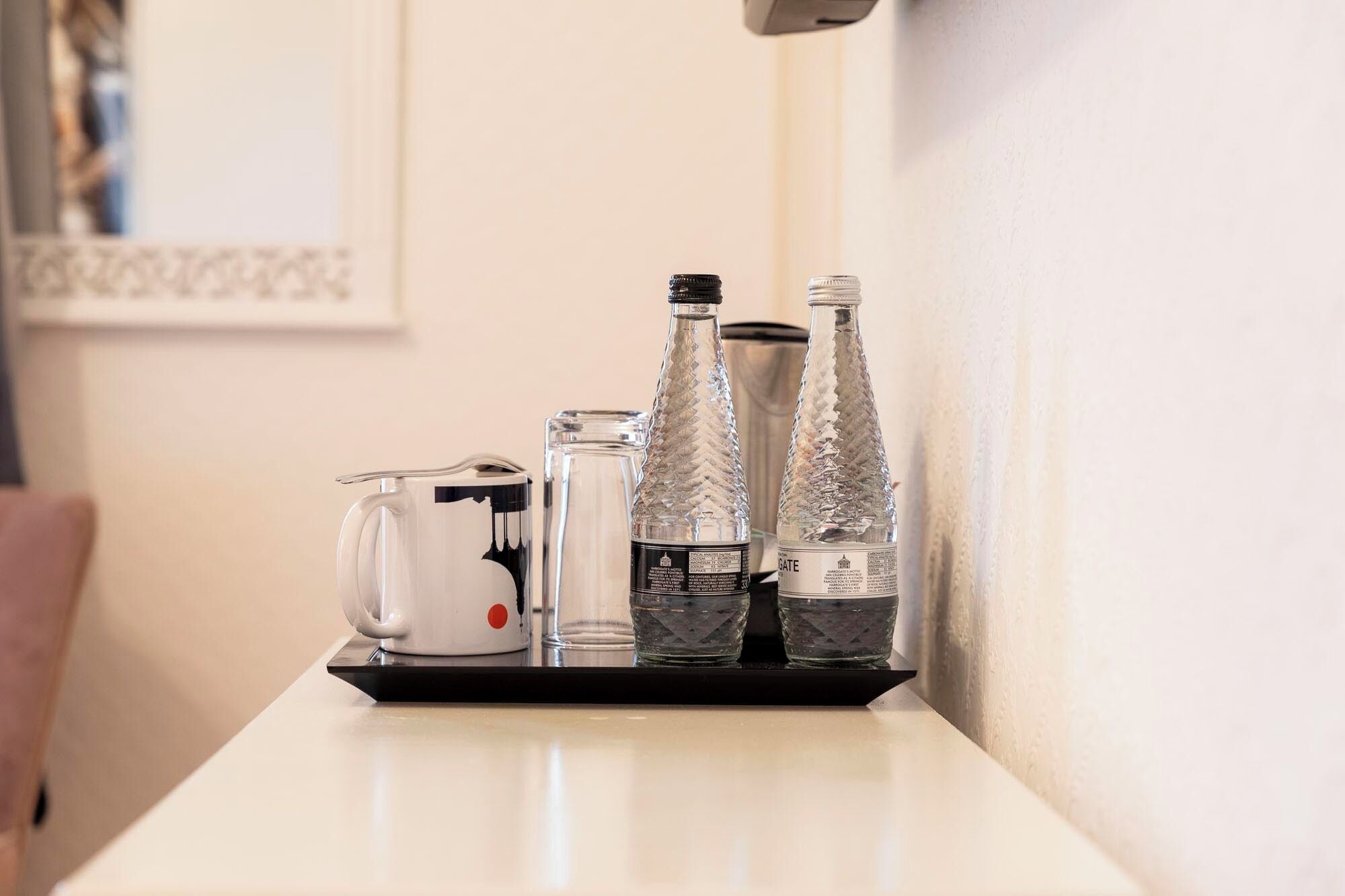 A white table with a black tray holding a white teapot, two clear glass bottles, and a glass.