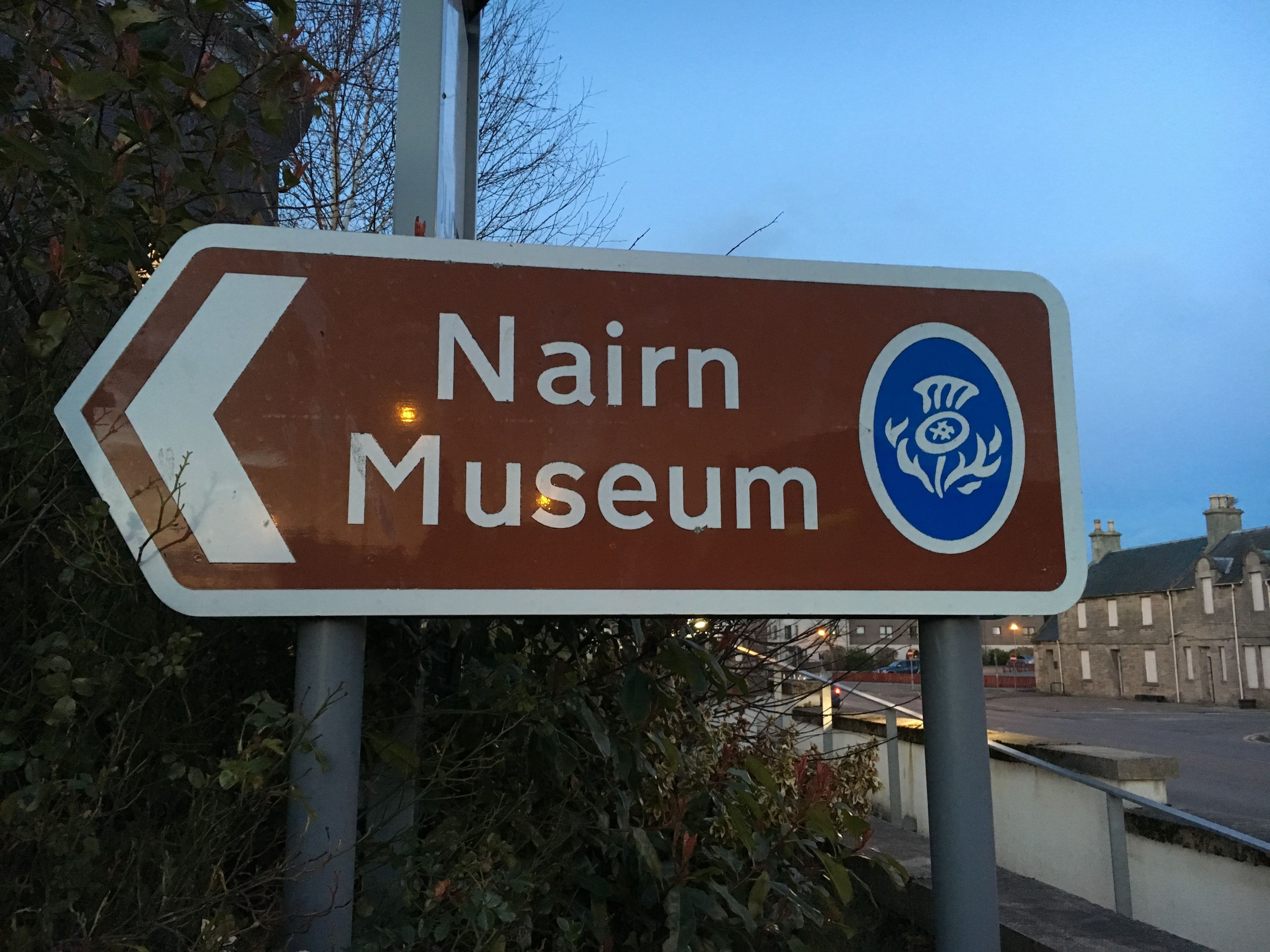 A directional sign for the Nairn Museum with a blue emblem.
