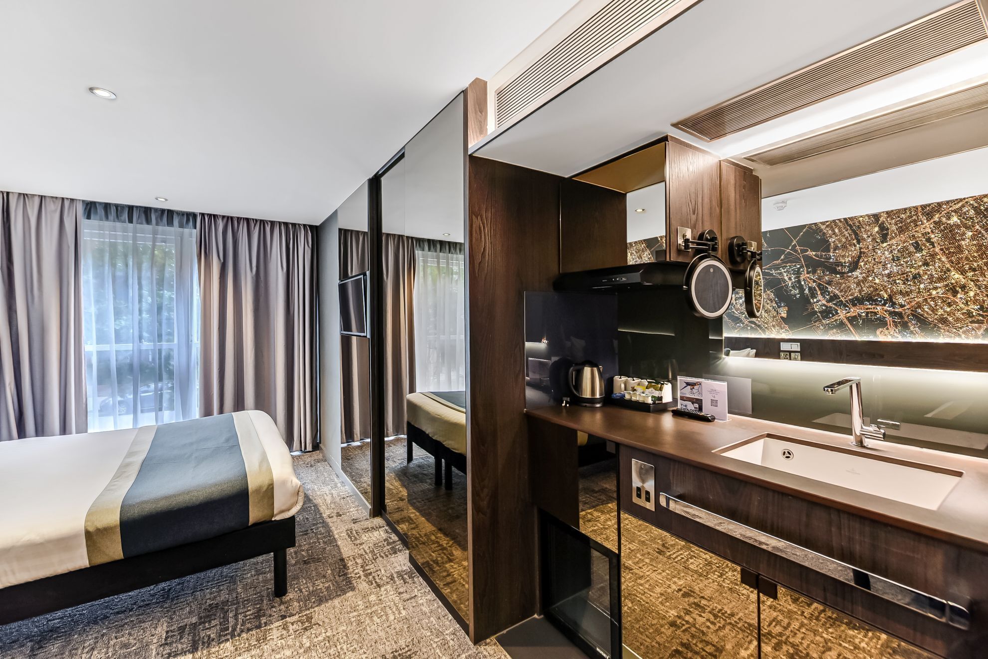Une chambre d’hôtel moderne équipée d’un lit, d’un bureau avec un lavabo, d’un miroir et d’une œuvre d’art représentant un paysage urbain.