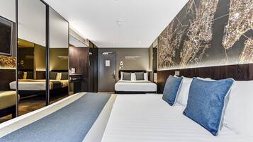A modern hotel room with a large bed, a desk, a mirror, and a cityscape artwork on the wall.