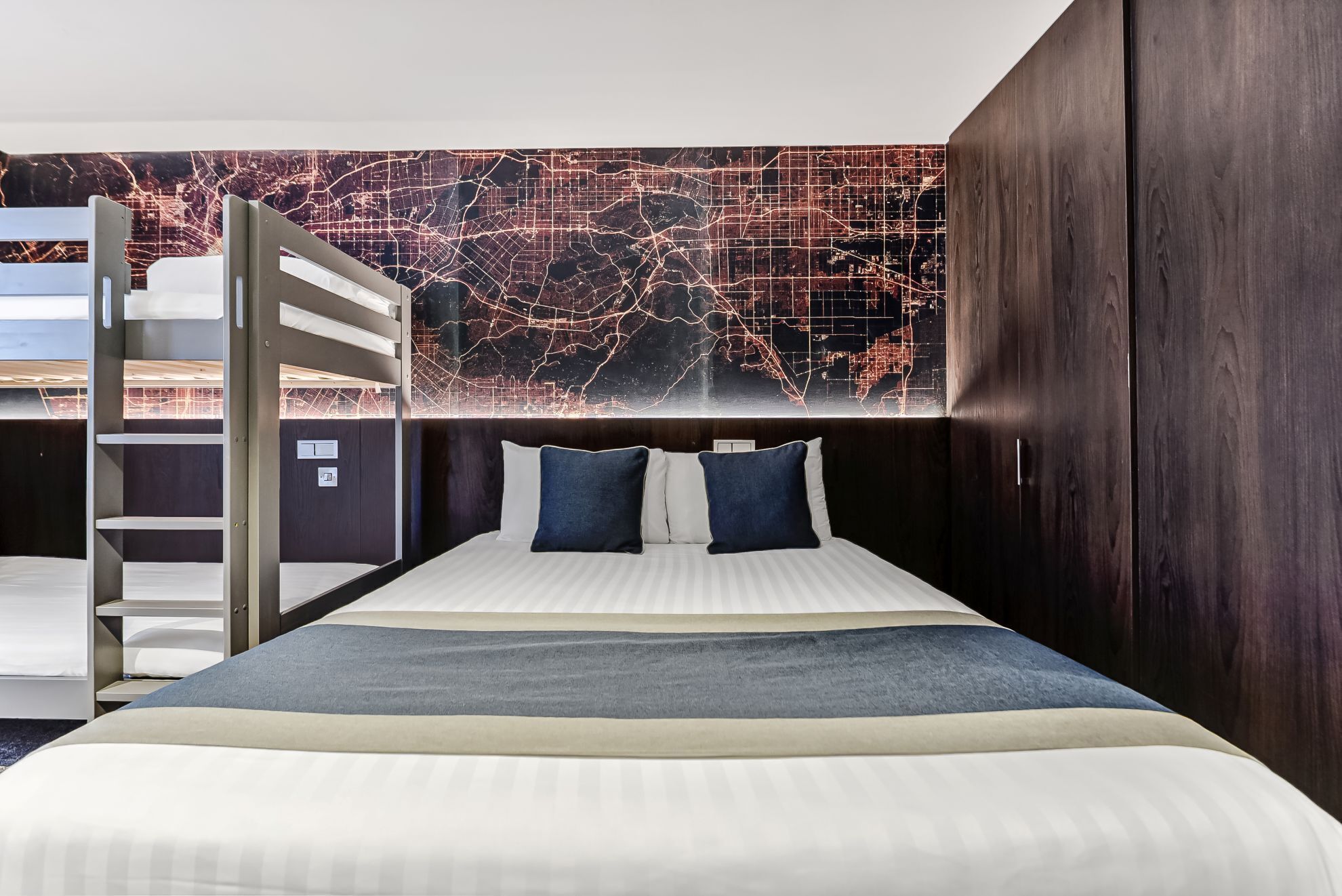 A modern hotel room with a bed, a map mural, a wooden wardrobe, and a bunk bed.