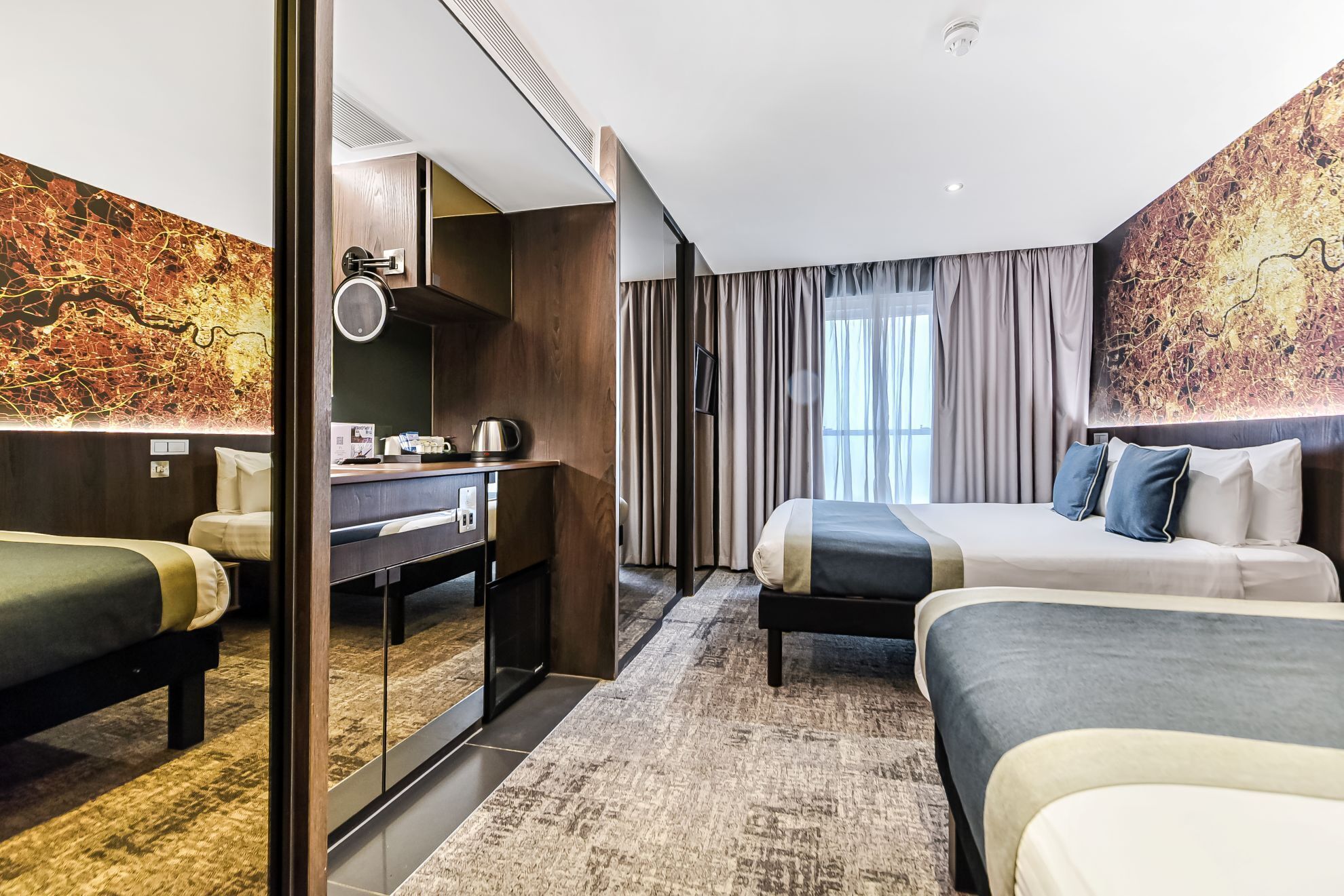 Ein Hotelzimmer mit Schreibtisch, zwei Betten, einem großen Fenster mit Vorhängen und einer Wand mit strukturierter Wandkunst.