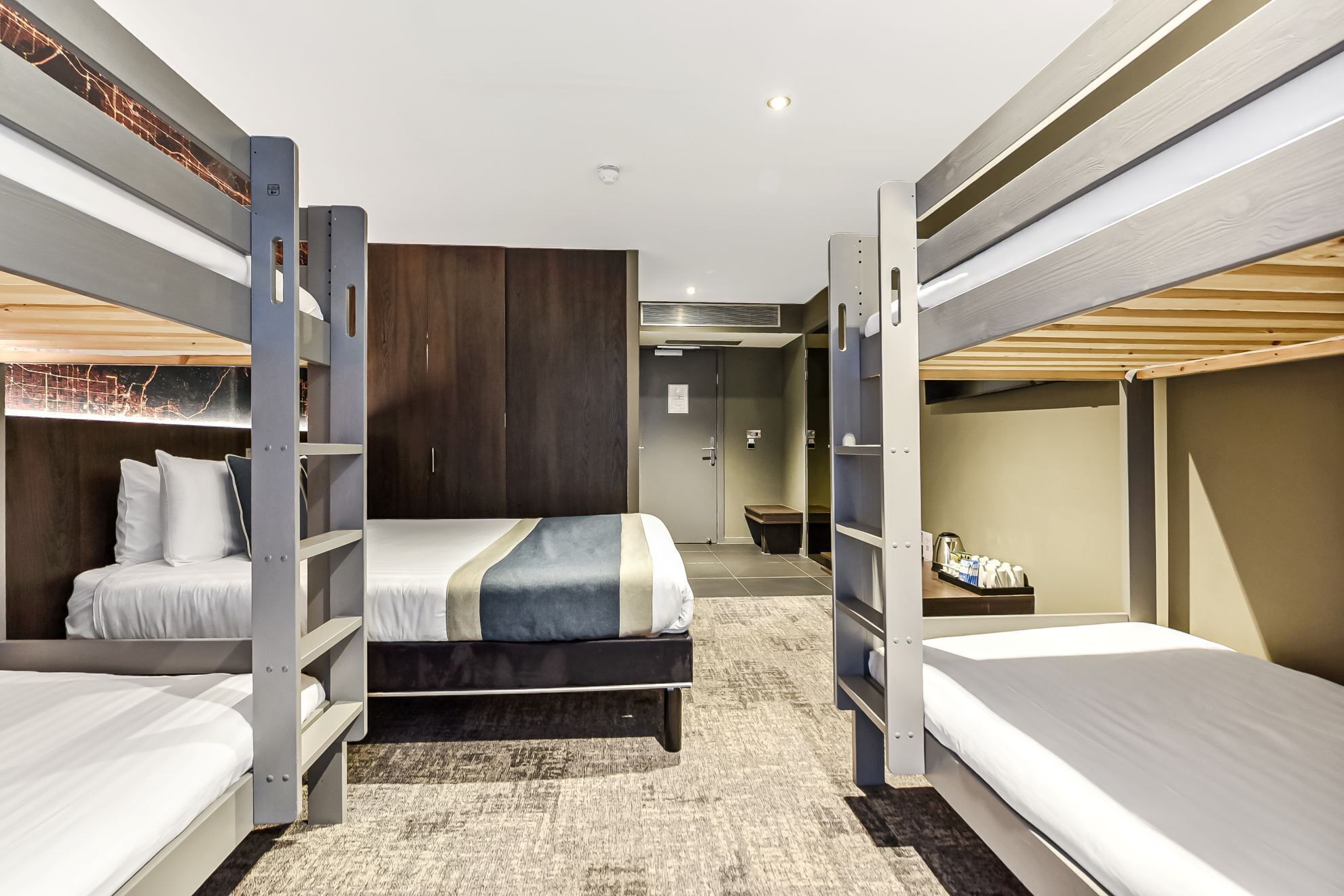 A hotel room with bunk beds, a wooden headboard, a small bathroom, and a kitchenette.