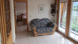 Ein Zimmer mit Holzboden, einem blau gemusterten Sofa, einer Holztür und einem Fenster mit Blick auf Grünflächen.