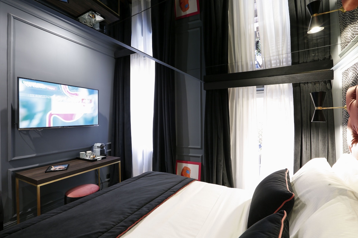 A modern hotel room with a bed, a desk with a computer, a TV, and a window with curtains.