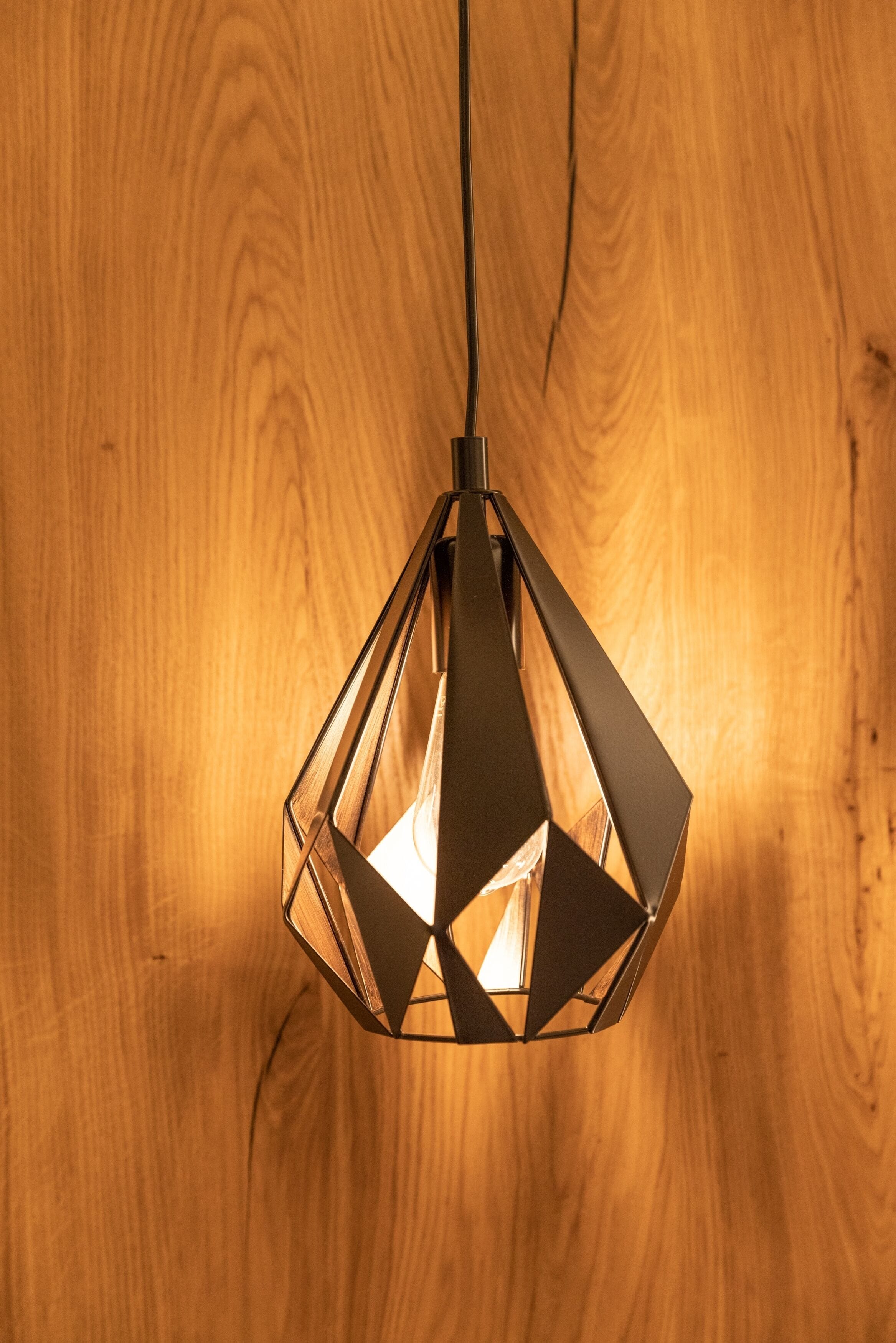 Lámpara colgante de diseño geométrico con bombilla visible, suspendida de un cordón sobre un fondo de madera.