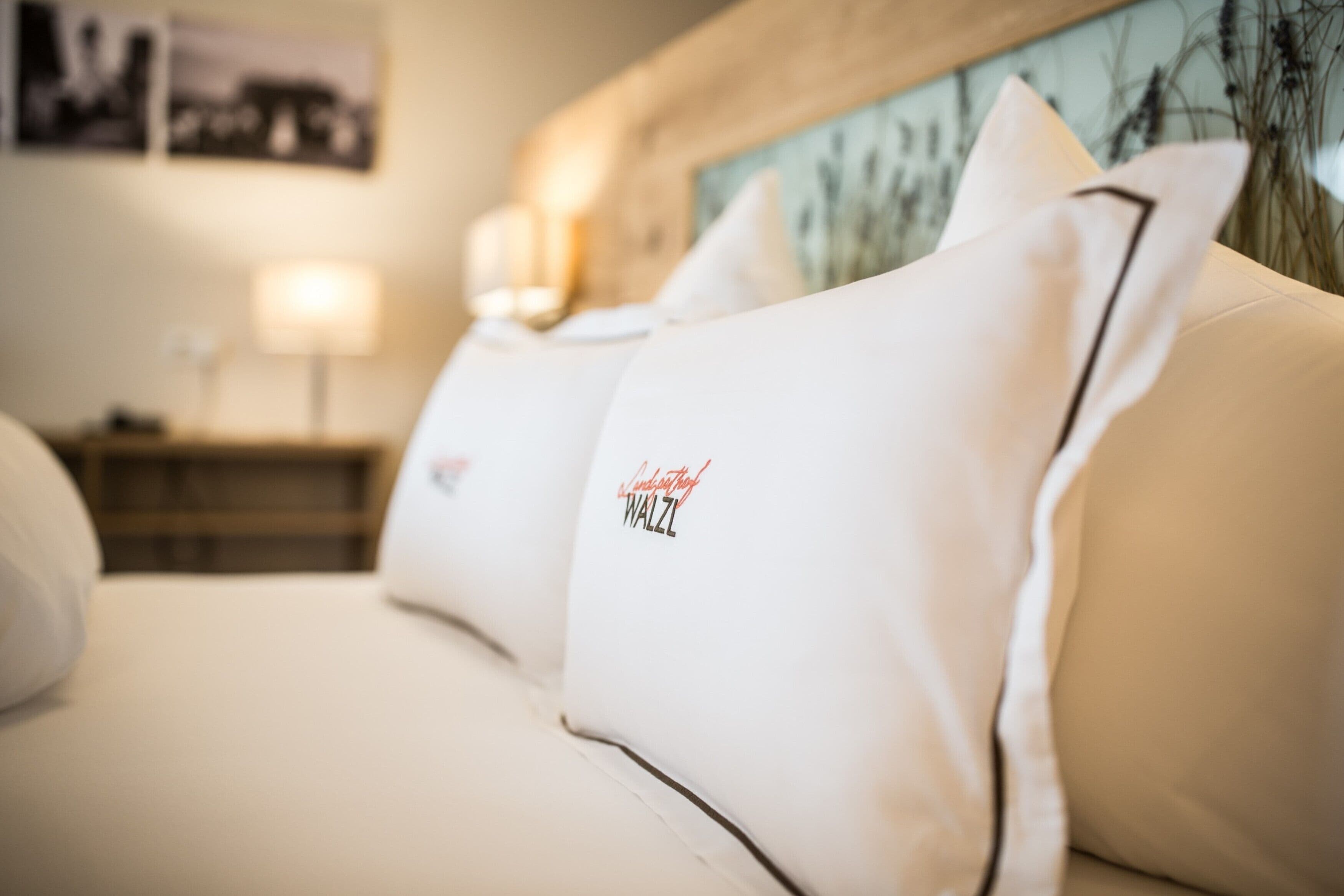 A hotel room with a neatly made bed, white pillows with a logo, a nightstand with a lamp, and a painting on the wall.