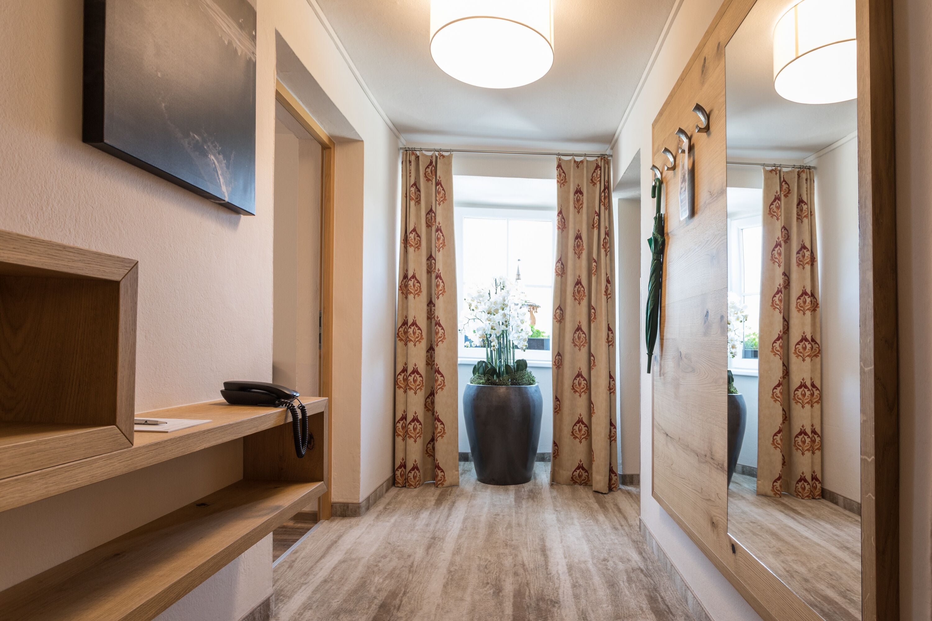 A hotel hallway with a wooden reception desk, a large potted plant, patterned curtains, and a mirror reflecting the interior.
