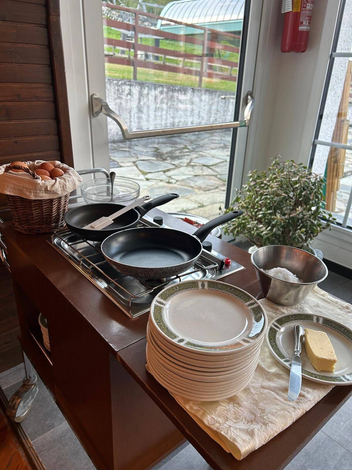 A kitchen counter with cooking utensils, a basket of eggs, a stack of plates, a bowl of salt, and a window with a view of a fenced area.