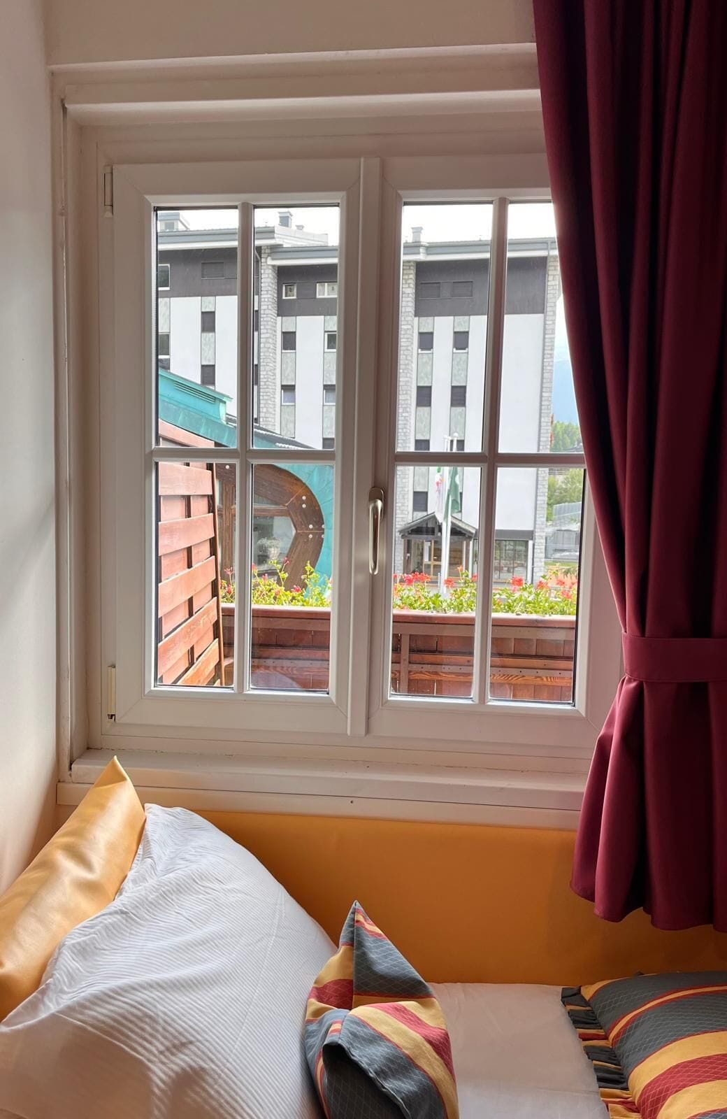A room with a bed, a window with curtains, and a view of buildings and a garden.