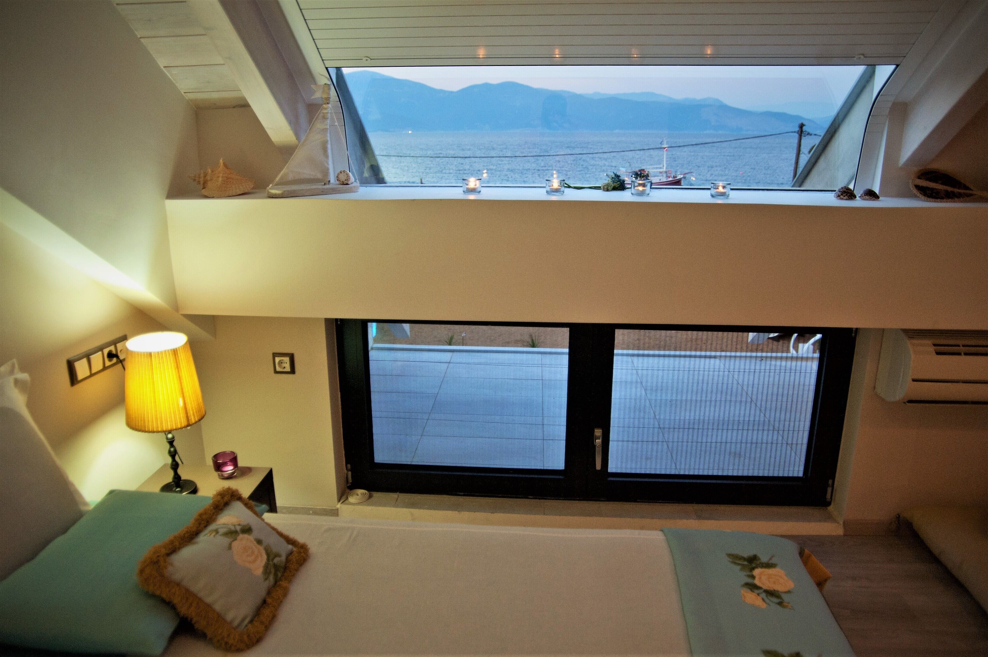 A bedroom with a skylight, a bed with pillows, a bedside table with a lamp, and a view of a dock and mountains.