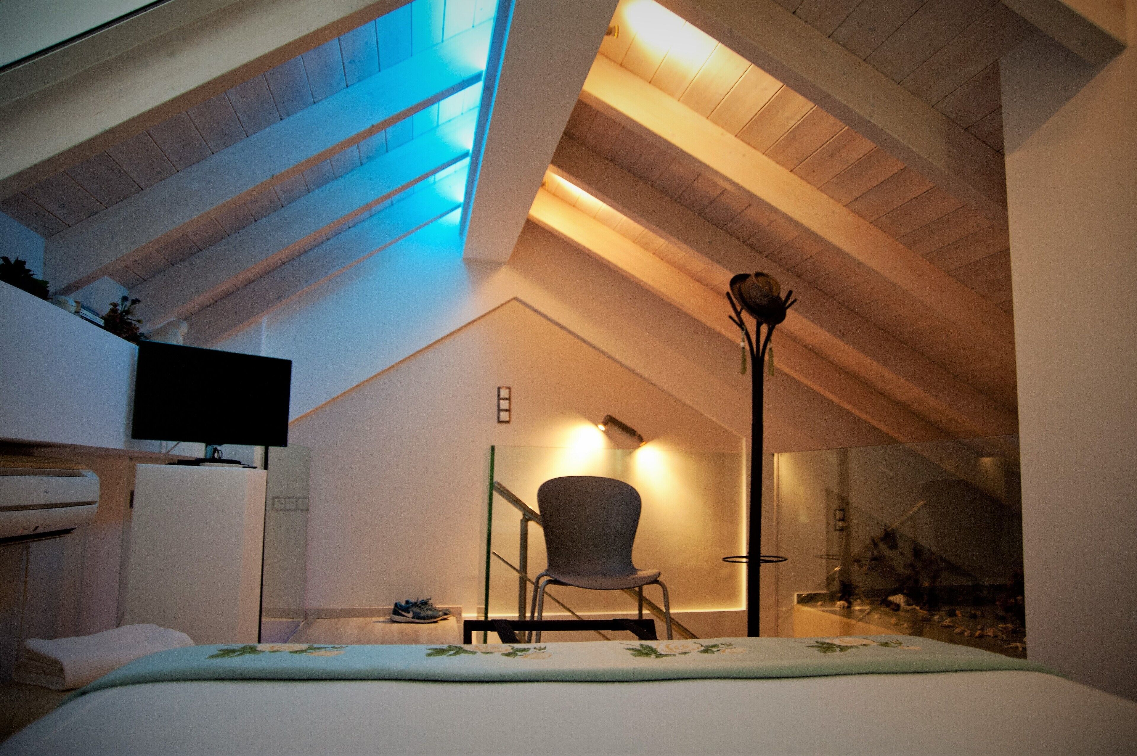Quarto moderno com teto inclinado, cama, poltrona, televisão e divisória de vidro.