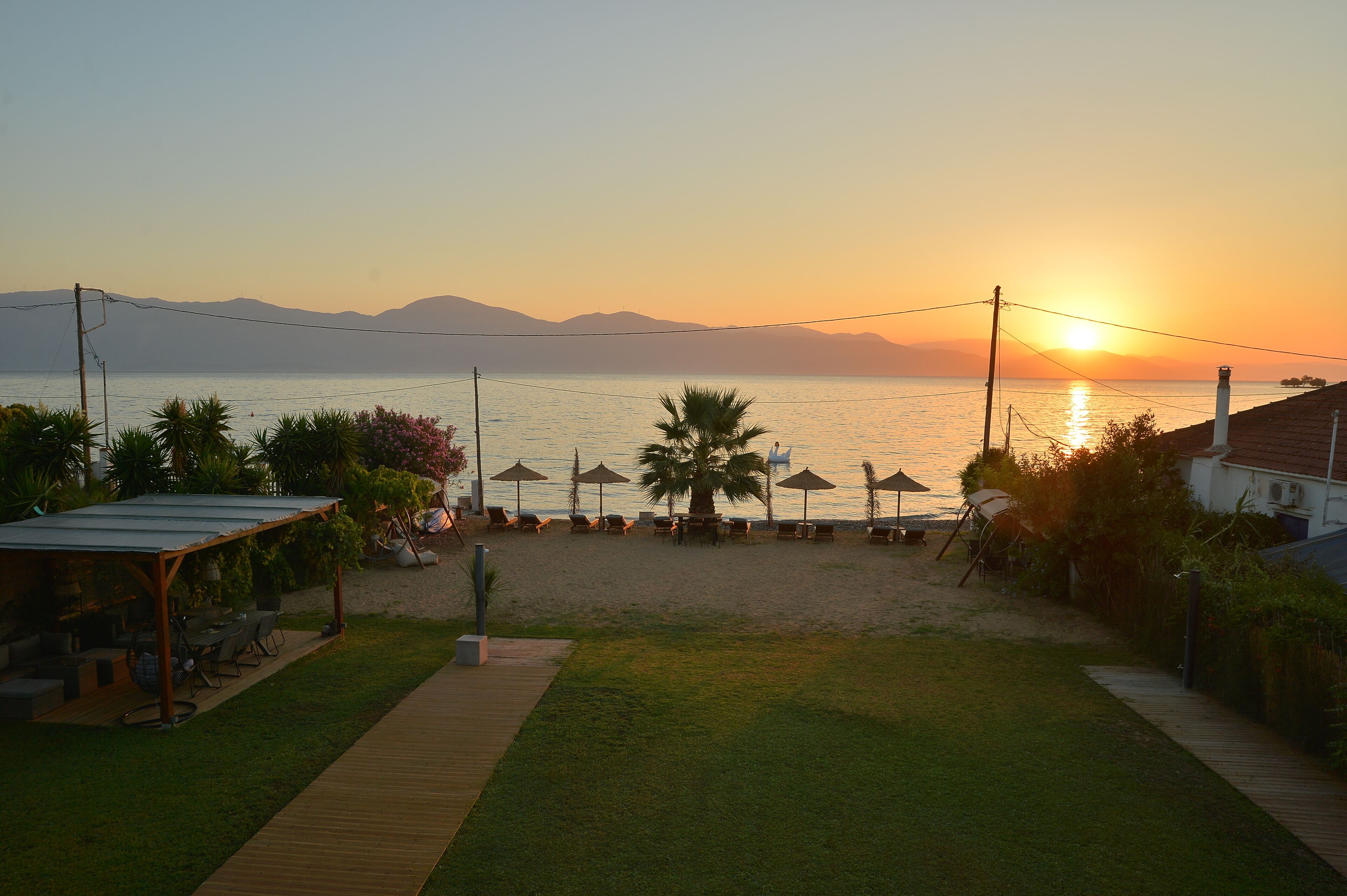 Terrassa al costat de la platja amb seients exteriors i ombrel·les, amb vista a un mar tranquil i muntanyes.