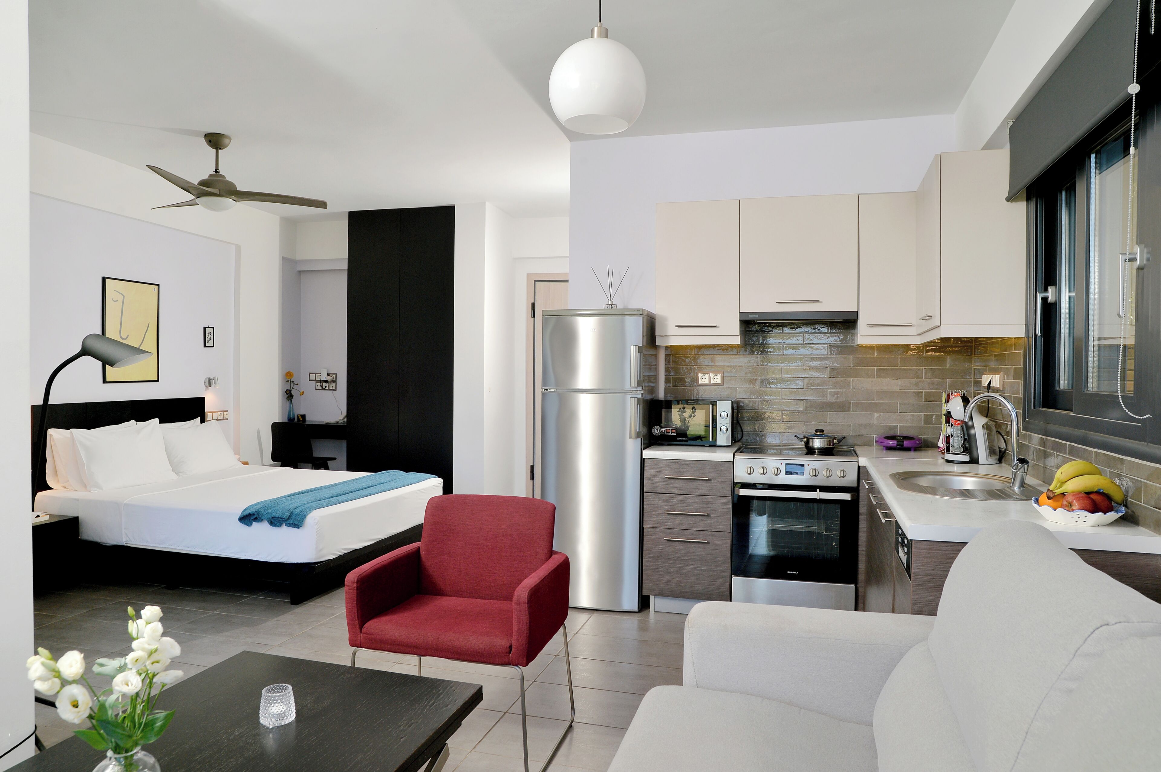 A compact hotel room with a bed, a red armchair, a white sofa, a kitchen area with modern appliances, and a ceiling fan.