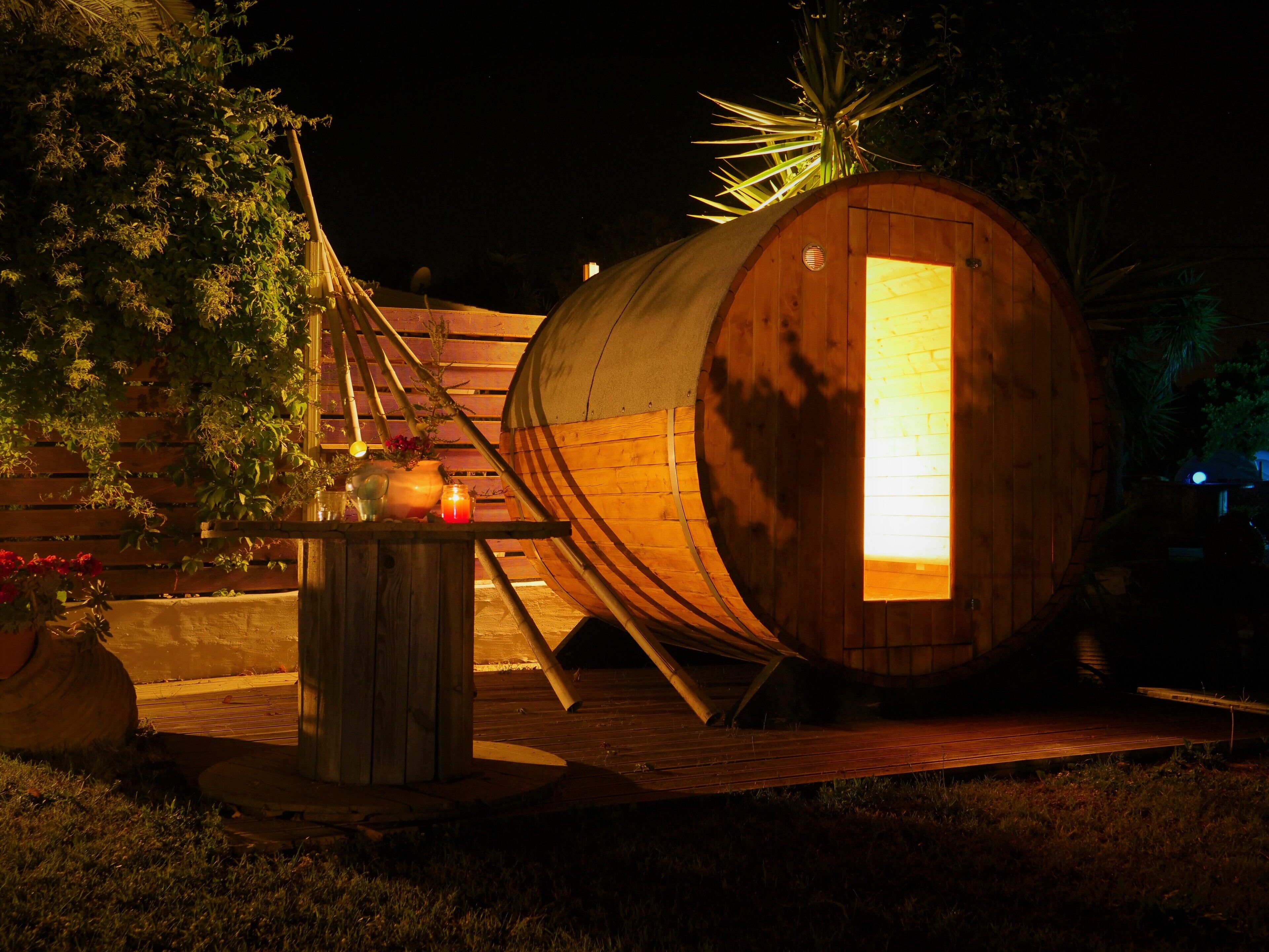 Una estructura en forma de barril de madera con interior iluminado, rodeada por una terraza de madera y macetas con plantas.