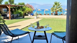Um pátio com duas cadeiras de descanso, uma mesa com prato de comida e bebidas, e vista para a praia e montanhas ao fundo.