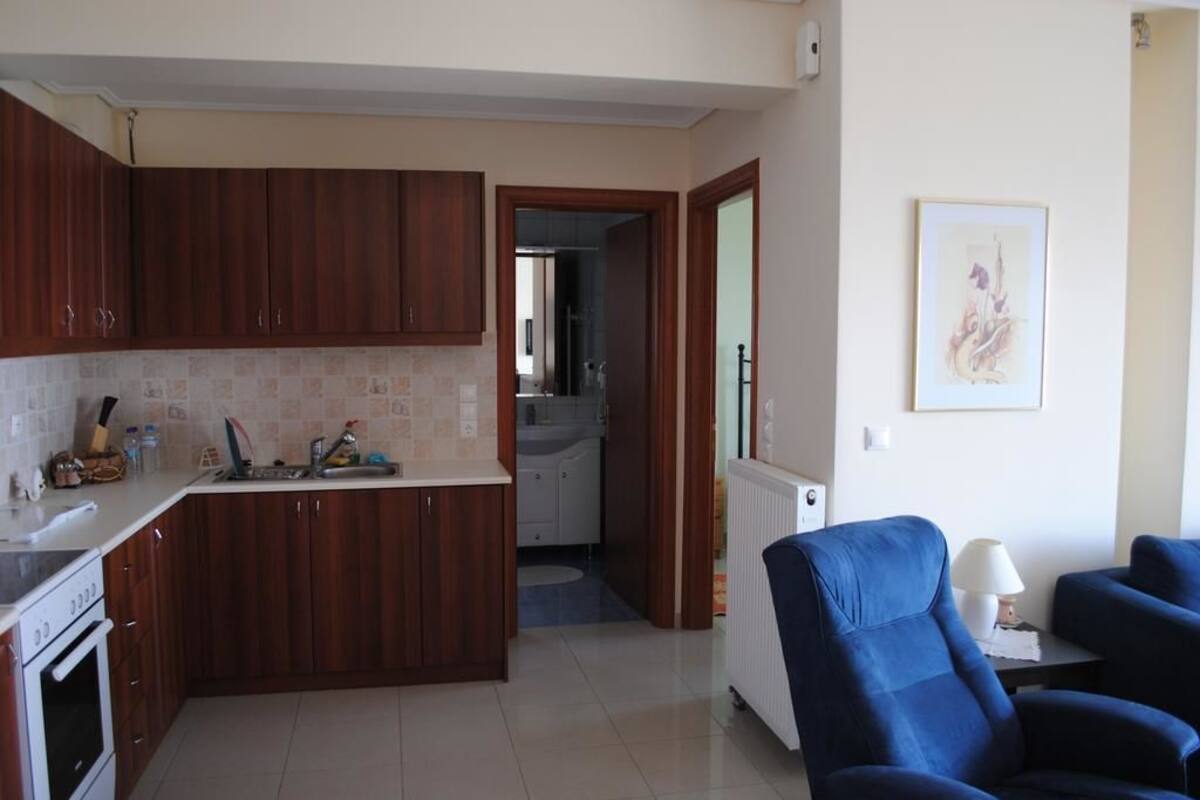 Una cocina con armarios de madera, una encimera blanca y un horno empotrado. En la zona de estar, hay una silla azul y una mesa con una lámpara.