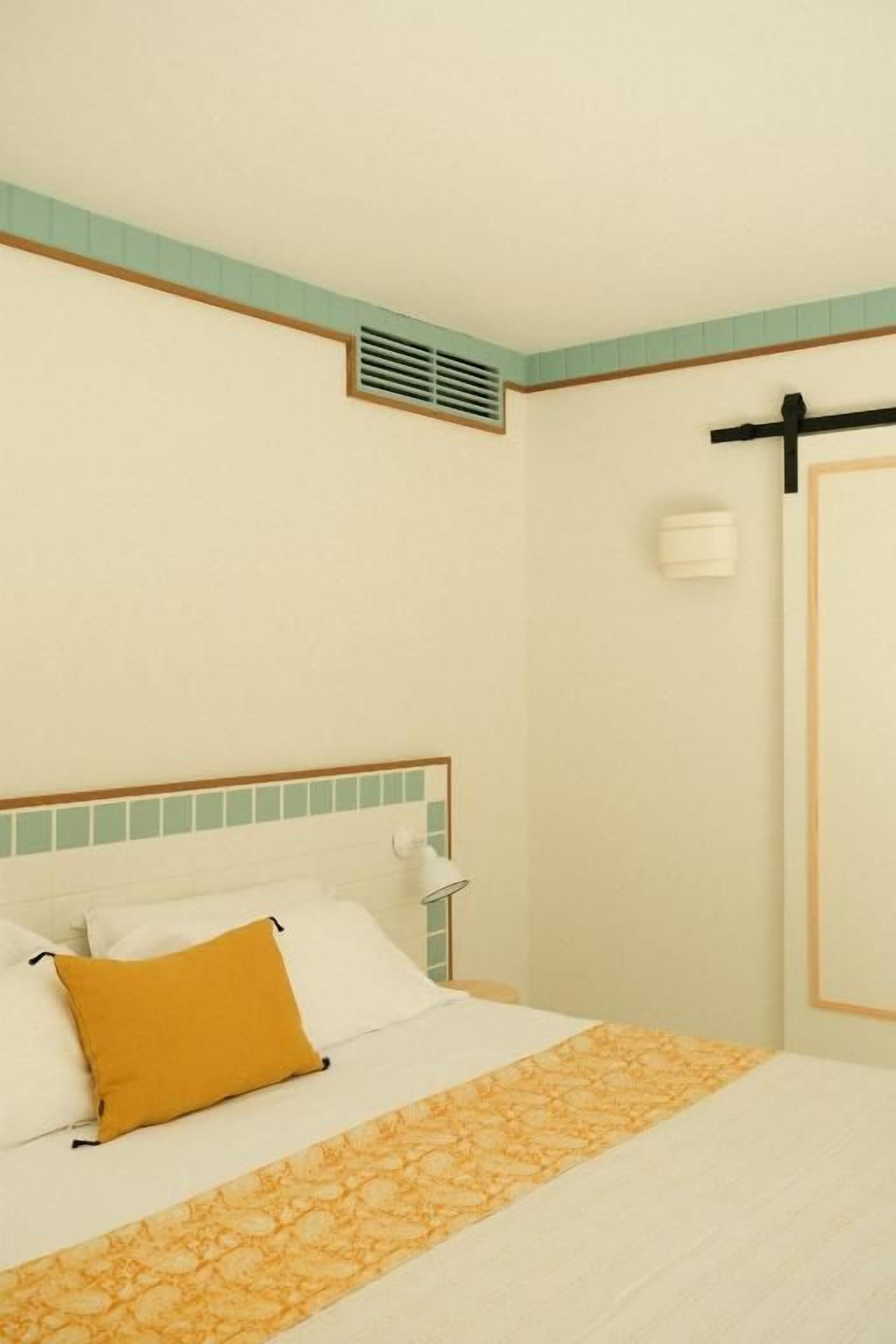 Une chambre d’hôtel avec un lit, une table de chevet, une lampe fixée au mur et une porte dotée d’une poignée.