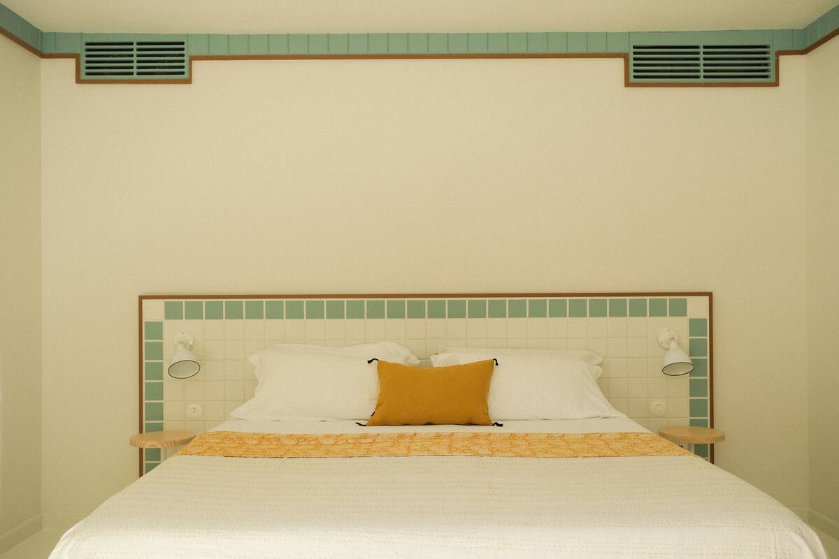 A neatly made bed with white and yellow bedding, a green-tiled headboard, and two wall-mounted light fixtures.