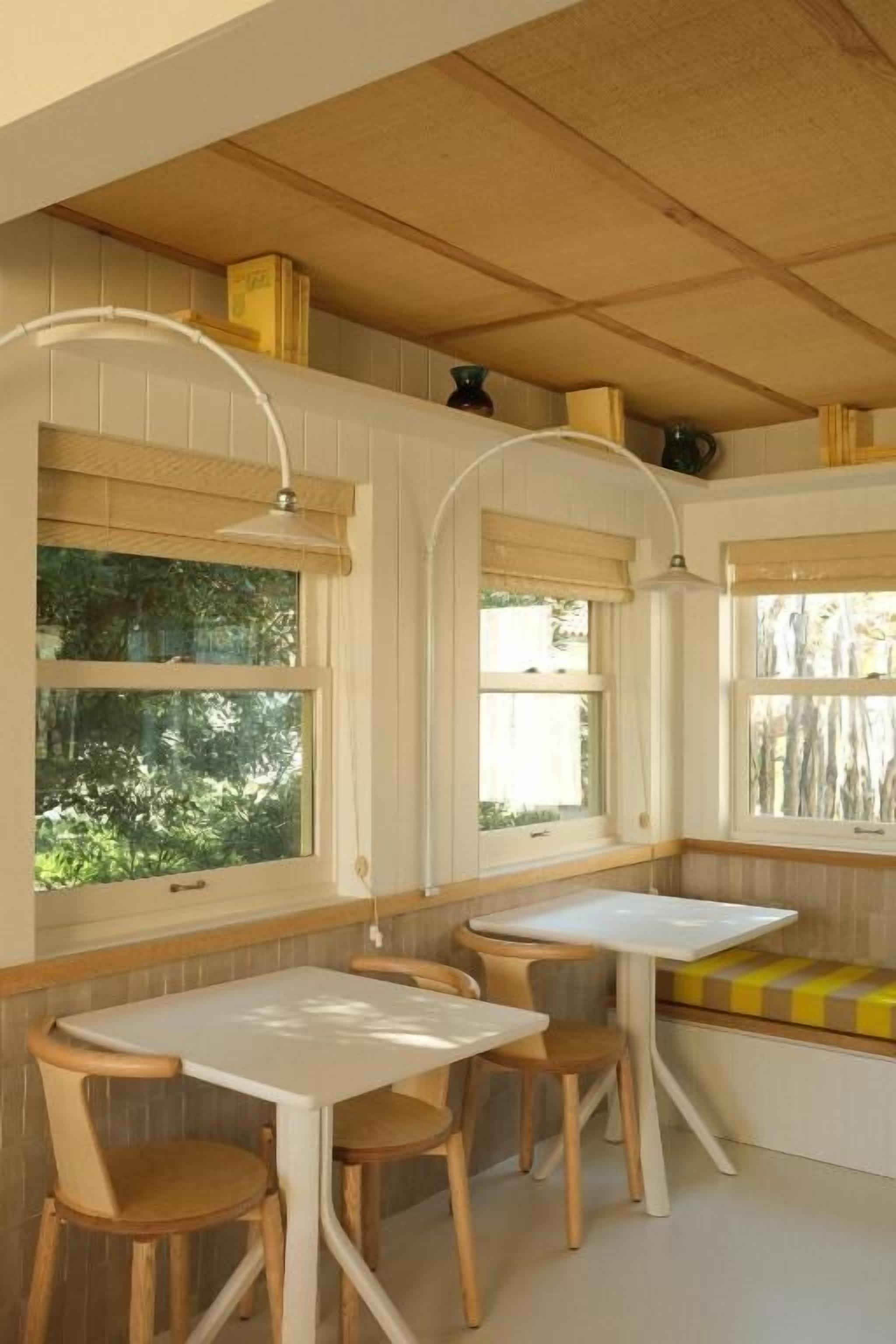 A room with a wooden ceiling, white walls, and a window seat with a bench. There are two white tables and four chairs.