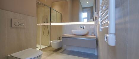 Un baño moderno con inodoro, bidé, ducha con cerramiento de vidrio y un espejo grande sobre el lavamanos.