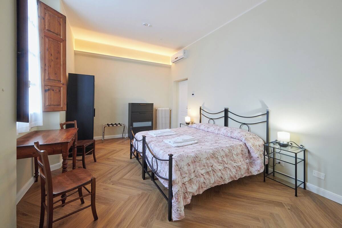 A bedroom with a bed, a desk, a chair, a radiator, and a small table with a lamp.