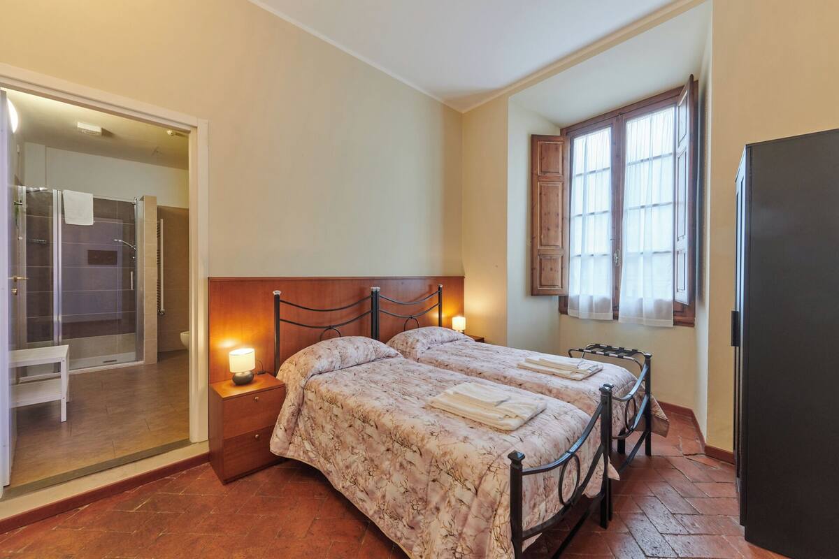 A bedroom with two beds, a wooden headboard, a nightstand with a lamp, a window with curtains, and a door leading to a bathroom.