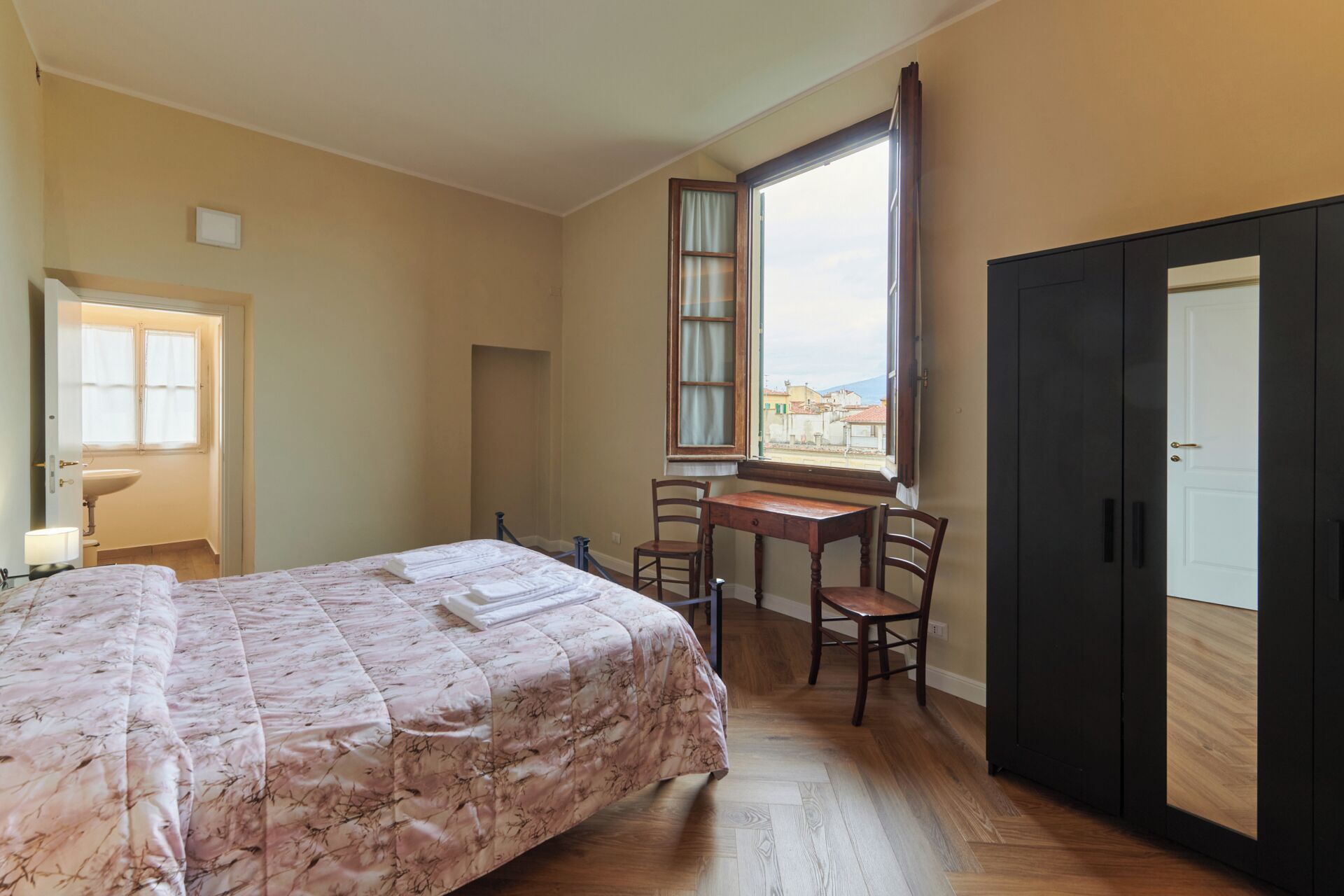 A bedroom with a bed, a desk, two chairs, a window, and a wardrobe.