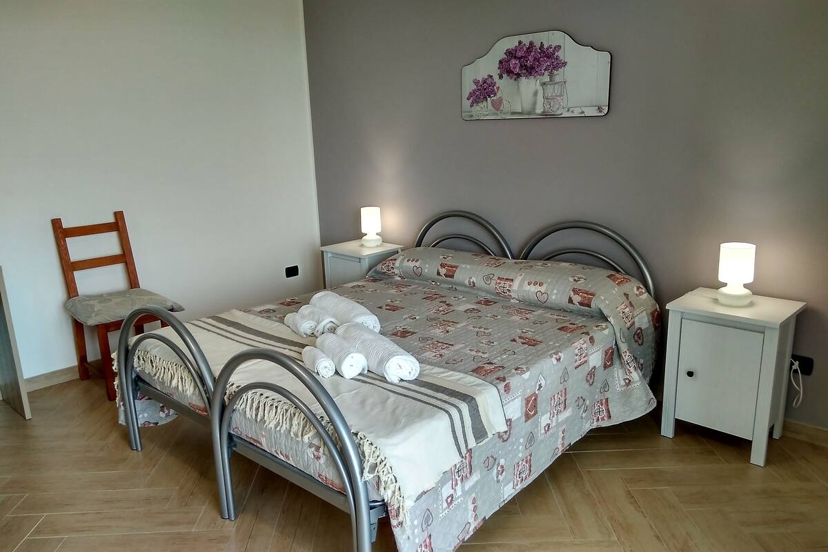A neatly made bed with a floral bedspread, two bedside tables with lamps, a wooden chair, and a wall-mounted decoration.