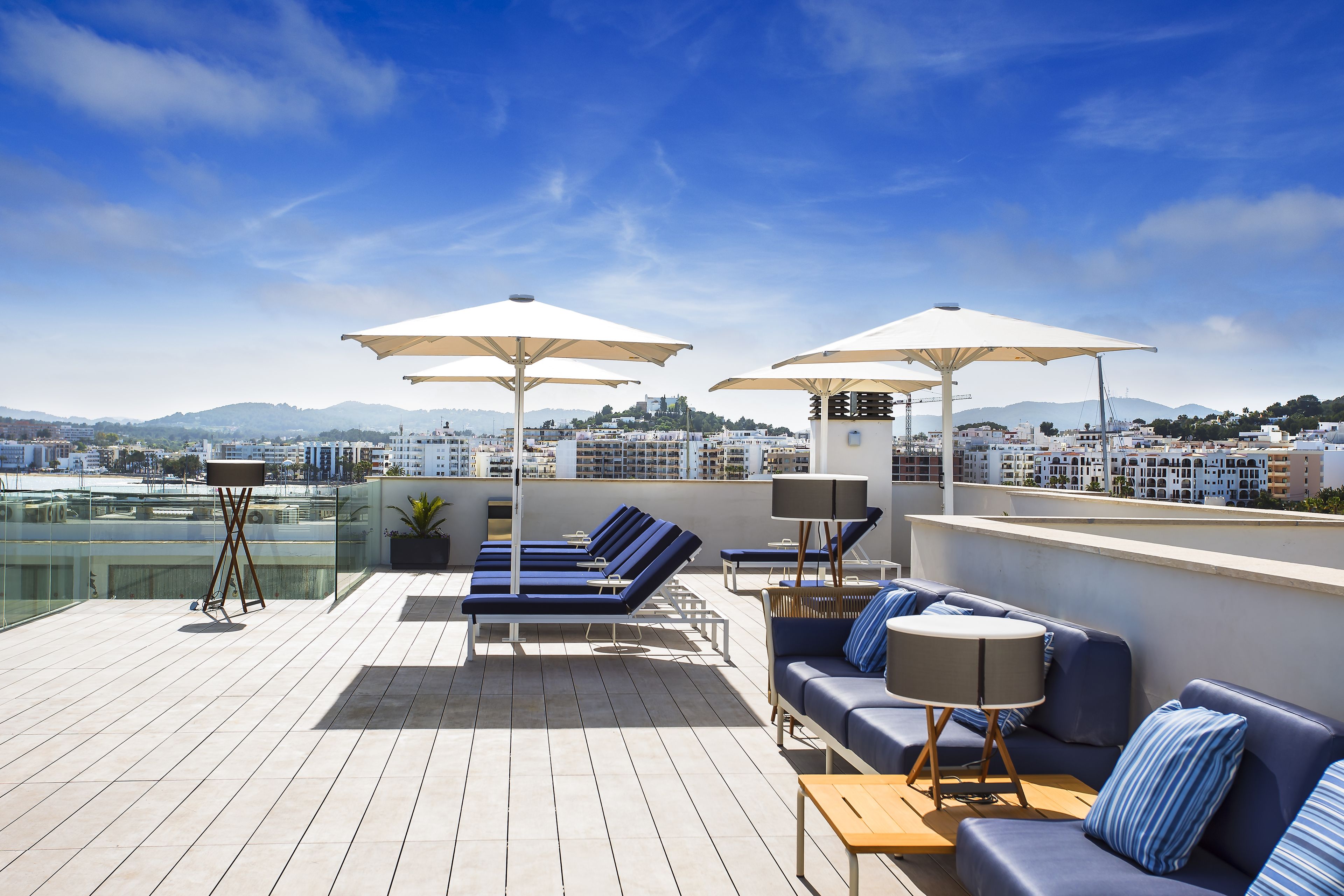Una terraza en la azotea con sillones de sol, un sofá y sombrillas, con vistas a un paisaje urbano y montañas al fondo.