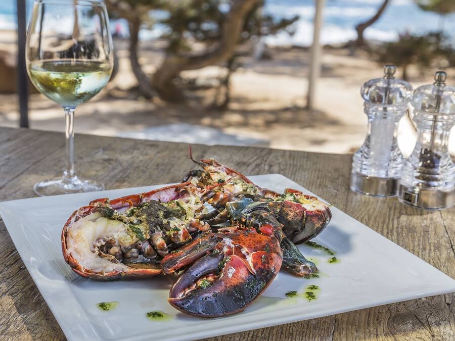 A lobster dish on a white plate with a glass of white wine and salt and pepper shakers on a wooden table.