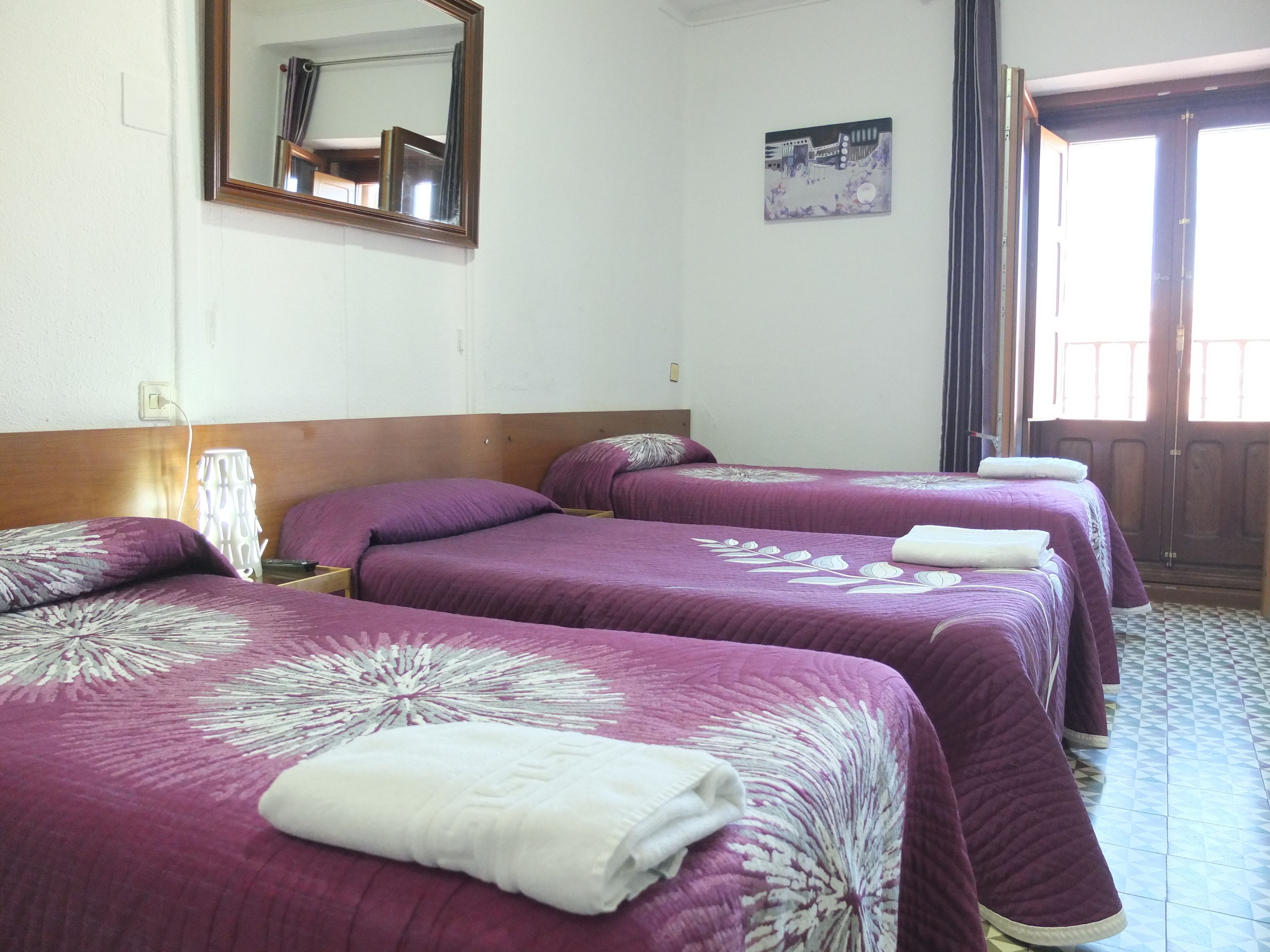 A hotel room with three beds, each with a purple bedspread and white pillows. There is a mirror on the wall, a painting, and a door with a view.