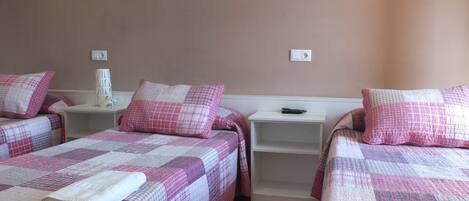 Una habitación con dos camas, cada una con una colcha y almohadas a cuadros rosa y blanco. Se observan mesitas de noche blancas, con un control remoto en una de ellas. Las paredes están pintadas de un tono melocotón claro, y hay dos interruptores de luz instalados en la pared.