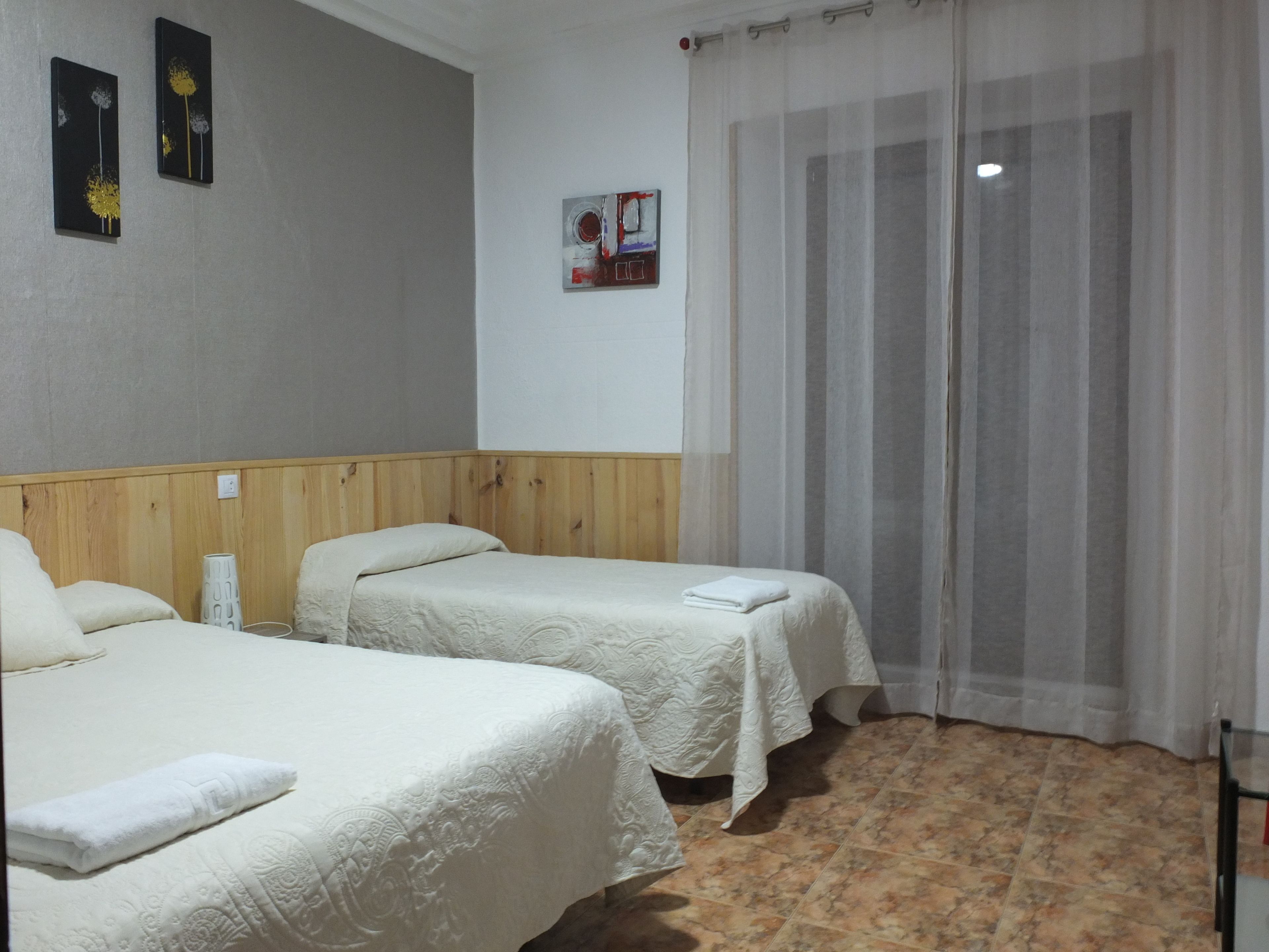 Habitación de hotel con dos camas, cabecero de madera, ventana con cortinas translúcidas y suelo de baldosas.