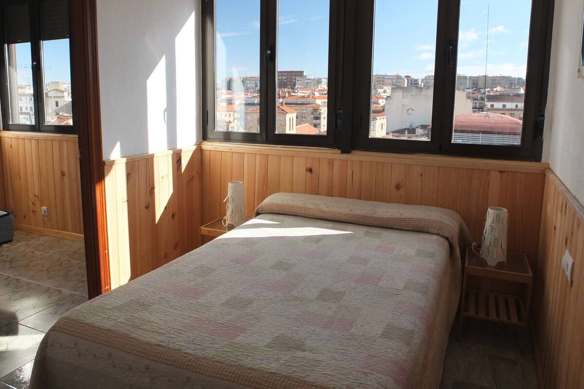 Une chambre avec une grande fenêtre, un lambris en bois, un lit recouvert d’une couvre-lit à motifs et une table de chevet avec une lampe.
