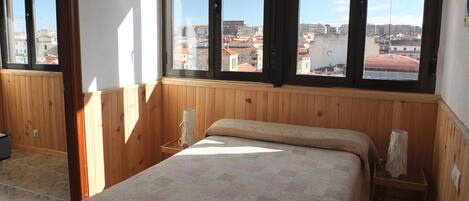 A bedroom with a large window, wooden paneling, a bed with a patterned bedspread, and a nightstand with a lamp.