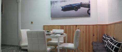A dining area with a white table, two white chairs, a wooden bench, and a large painting of a boat on the wall.