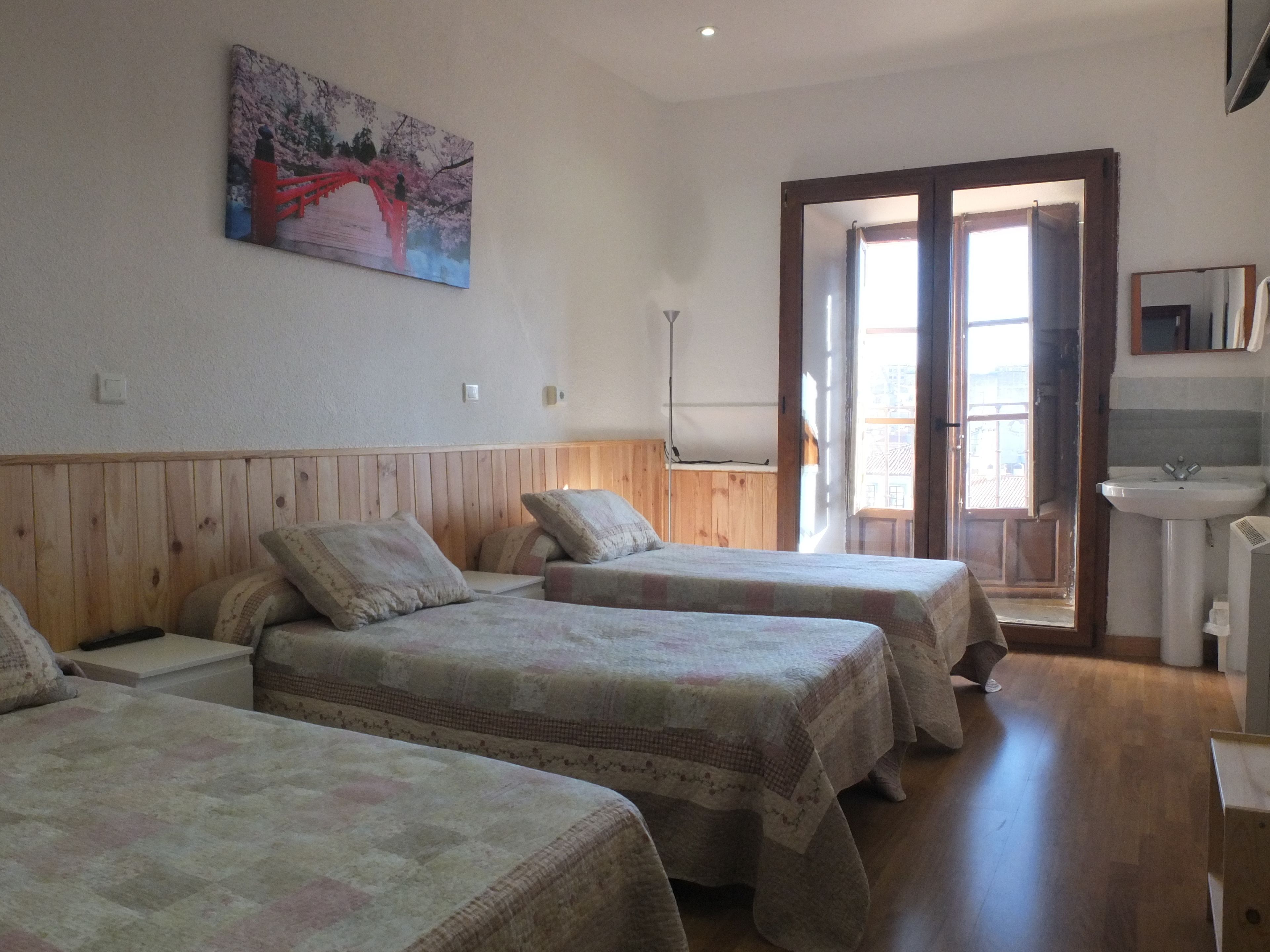 A room with three beds, a wooden headboard, a painting on the wall, a window with a view, and a bathroom with a sink and mirror.