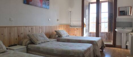 A room with three beds, a wooden headboard, a painting on the wall, a window with a view, and a bathroom with a sink and mirror.